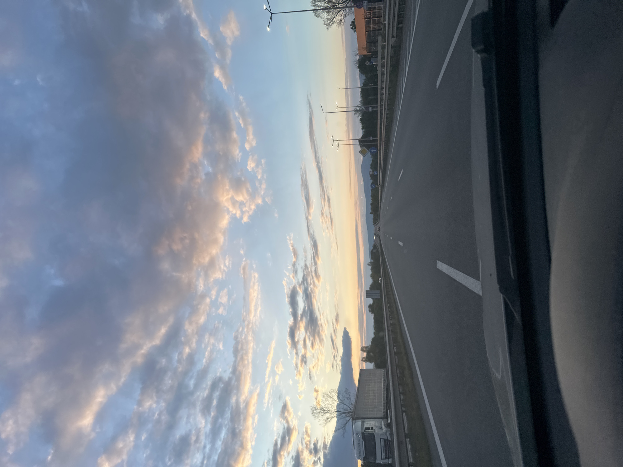 This image depicts a highway scene at either sunrise or sunset, capturing a vast expanse of sky and road from the perspective of a vehicle. The sky is a prominent feature, transitioning from clear blue at the top to warm hues of orange, pink, and yellow near the horizon. Numerous clouds are scattered across the sky; some appear dark and dense, while others are brightly illuminated by the low-angled sun, creating strong atmospheric contrast.
A multi-lane highway stretches into the distance, marked with distinct white lines and bordered by a barrier. Modern streetlights, some of which are illuminated, line the road. To the left, trees and distant, softly outlined mountain ranges are visible, blending with the horizon. These mountains are consistent with the provided geographical context of Sofia, Bulgaria. Road signs, including a yellow diamond-shaped warning sign and a round blue mandatory direction sign, are present along the roadside.
On the right side of the highway, a white semi-trailer truck is visible, positioned on the shoulder or a parallel lane. Parts of the vehicle from which the photo was taken are visible on the right, framing the scene. The overall impression is one of tranquil travel on an open road during twilight hours. No specific human figures or activities are discernable, and no legible text appears on any signs or objects within the frame.