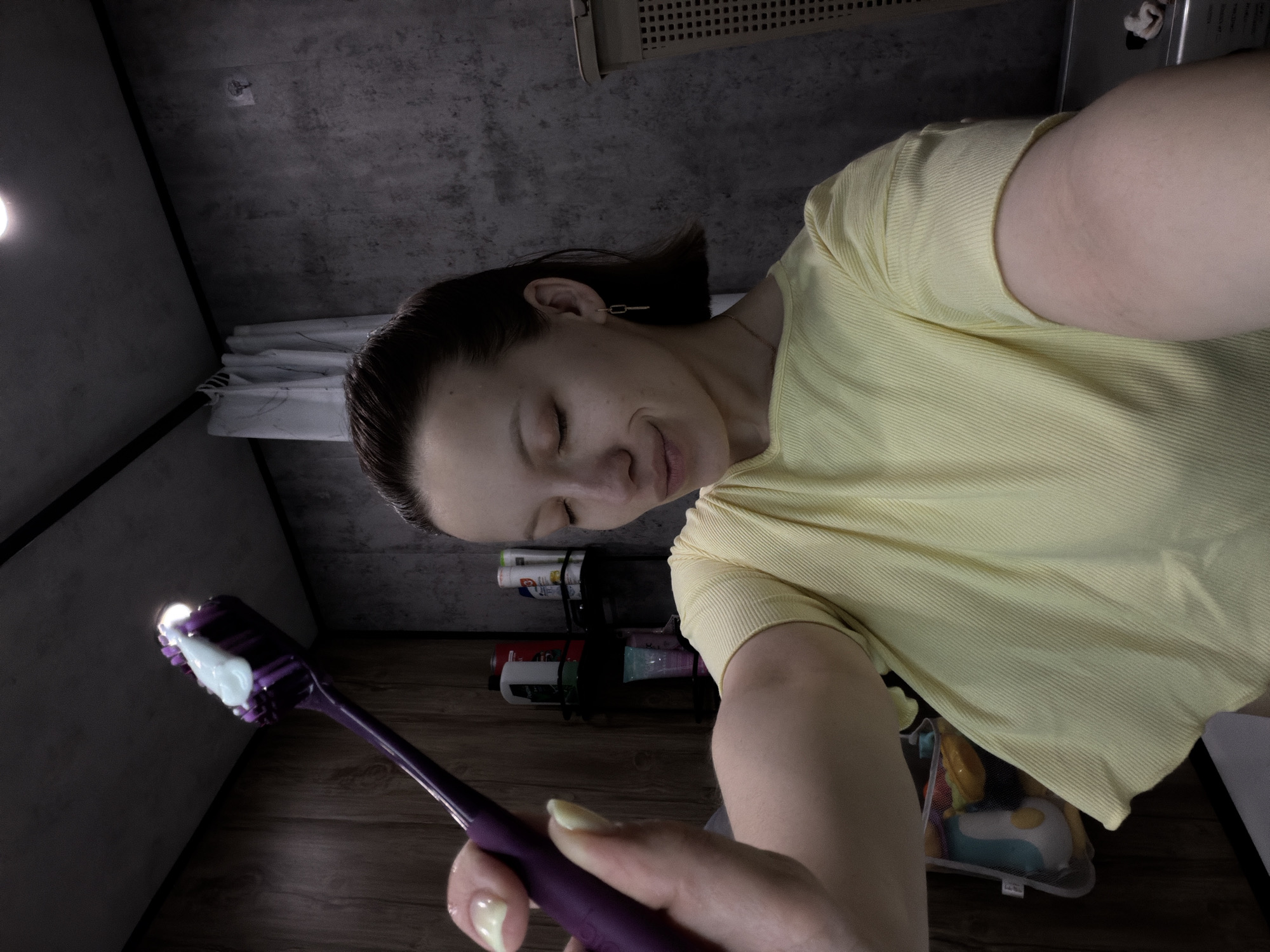 A woman is holding a purple toothbrush with white toothpaste in a bathroom. The bathroom has gray walls and wooden flooring. A shelf holding toiletries is visible in the background. The woman is wearing a yellow shirt. She has short brown hair and is wearing earrings. The lighting is dim, possibly from an overhead light. There is no evidence to suggest this image was taken in Reshetnikovo, Russia.