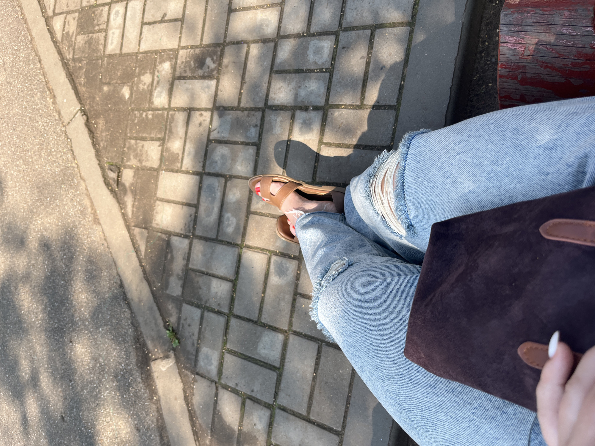 The image shows a close-up, overhead view of a person's legs and feet, wearing ripped light-wash denim jeans and tan sandals. The person is holding a dark brown bag in their lap. The scene appears to be outdoors, with a patterned paved surface and asphalt visible. Sunlight casts shadows across the ground. The location is identified as Chaikovsky, Russia.