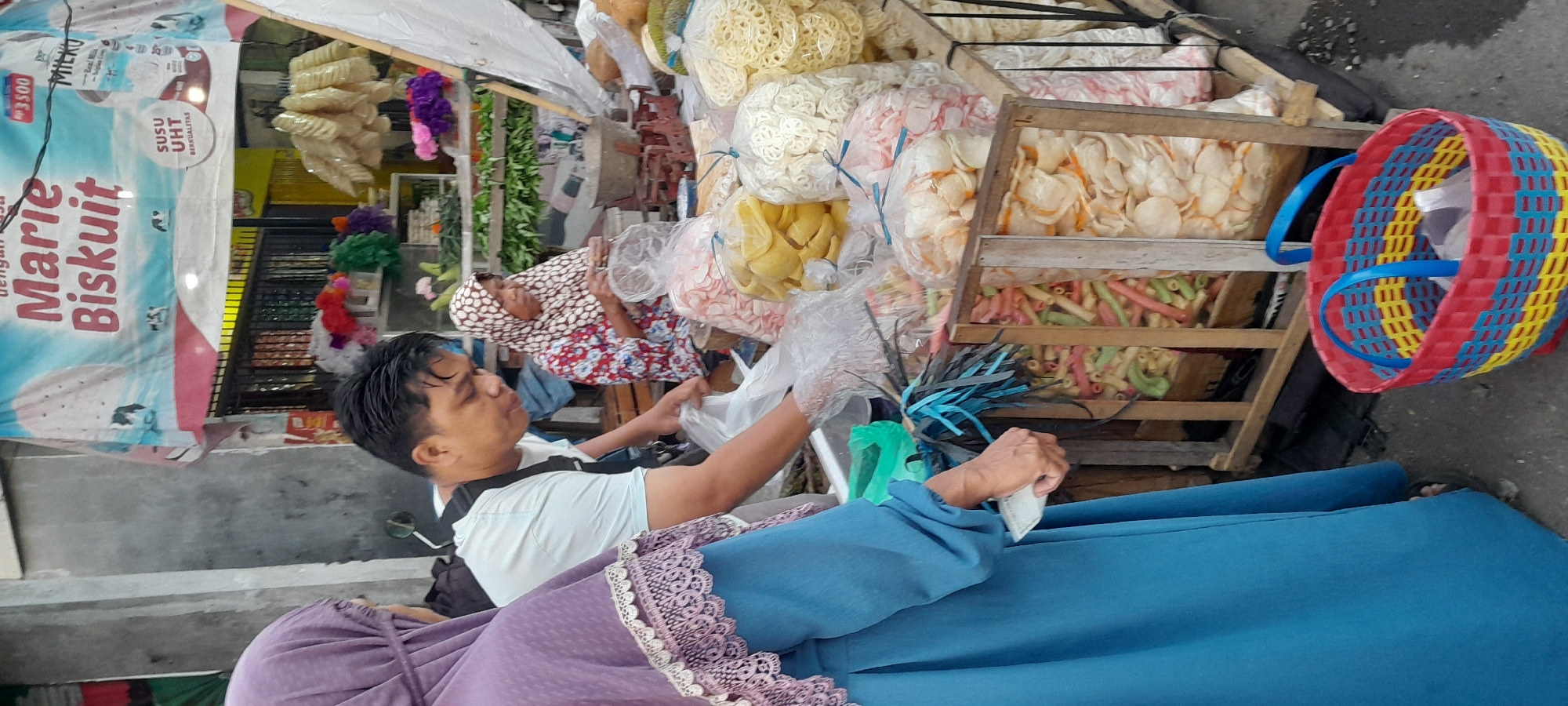 The image depicts a street market scene in Rogojampi, Indonesia, focusing on a stall selling various types of snacks and biscuits. In the foreground, a person wearing a blue hijab and a blue dress is partially visible on the left, holding a white paper or receipt in their gloved hand. To their right, a man wearing a light blue t-shirt and a black strap across his chest is engaged in handling plastic bags filled with snacks. He is looking down at the bags, suggesting an interaction with a customer or preparing an order. Behind them, an older woman wearing a patterned hijab and a red and white floral shirt is seated, also appearing to be involved in packaging or serving snacks.
The stall itself displays an array of colorful snacks, primarily crackers and puffed snacks, packaged in clear plastic bags. These bags are arranged in wooden crates and on shelves. Notably, a prominent banner in the background advertises "Marie Biskuit," indicating a specific brand being sold. There are also colorful decorative items, possibly artificial flowers, displayed on the stall. The environment appears to be outdoors, with a concrete or asphalt ground surface visible. The overall impression is one of a typical, bustling local market.