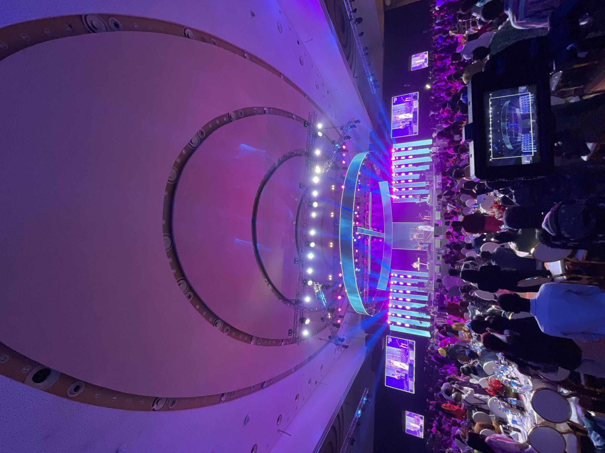 The image captures a large event space in Unknown, Unknown. A stage with a circular lighting rig is the focal point, illuminated with bright white and blue lights. Large screens flank the stage, displaying content to the audience. Attendees sit at tables, their backs facing the camera, suggesting a seated event like a gala or conference. The room is brightly lit with a purple hue, emanating from the stage lights and ceiling features. The ceiling has concentric circles, enhancing the architectural design of the event space. The image is taken from an elevated perspective, likely from a balcony or raised platform.