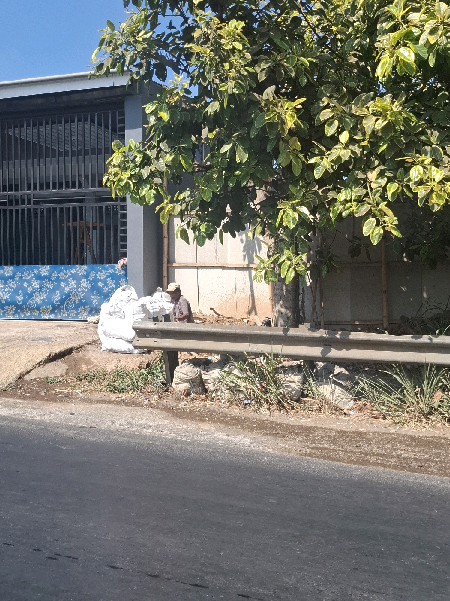 In the city of Cijalingan, Indonesia, a man wearing a light-colored cap and a dark t-shirt sits on the ground beside a stack of white sacks. He is positioned to the right of a large blue mattress with a white floral pattern, which leans against a building with a dark metal grate. The building is adjacent to a tree with lush green and yellow leaves. A metal guardrail runs along the edge of the road, separating it from the area where the man and the sacks are located. The ground is a mix of dirt, gravel, and sparse vegetation.