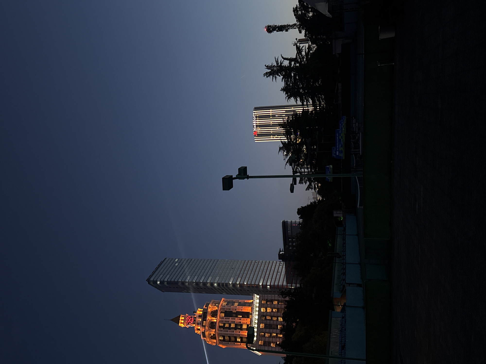 The image shows a nighttime view of several buildings in Batumi, Georgia, under a dark blue sky transitioning to lighter blue near the horizon. The building on the left is a tall structure with an ornate, brightly lit crown and a modern, glass-fronted tower. The Marriott hotel building is visible further to the right, distinguished by its illuminated "Marriott" sign. The scene includes streetlights and trees silhouetted against the sky. A tennis court is partially visible on the right.