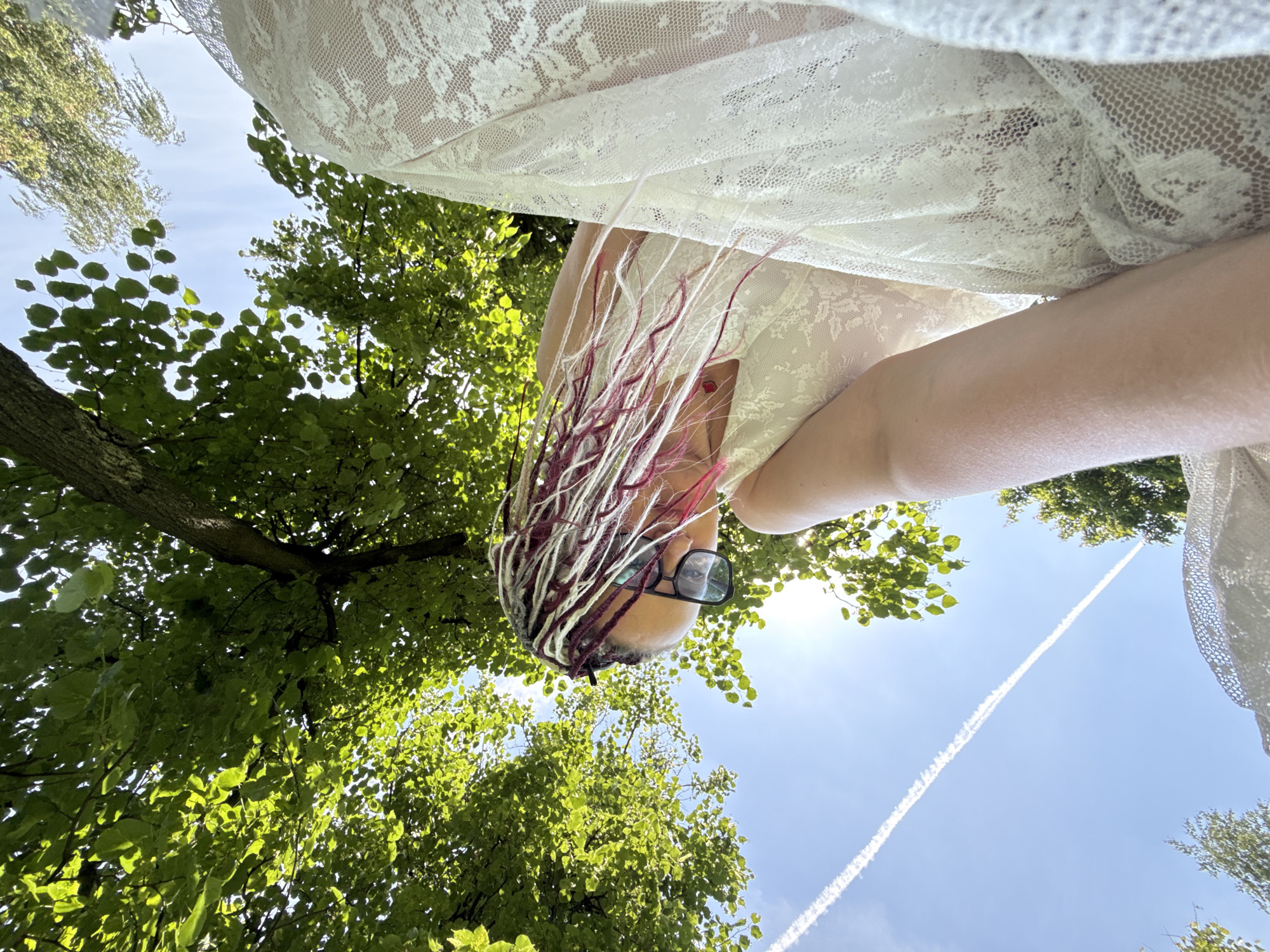 The image captures an outdoor scene, reportedly located in the city of Moscow, Russia, depicting an individual in a park or wooded area. The camera angle is low, looking upward. A person, visible from the upper chest to the head in the lower-right quadrant, is looking skyward. This individual has numerous dreadlock-like hair strands in a mix of white/blonde and maroon/red colors and wears dark-framed eyeglasses. They are dressed in a light-colored, possibly white, lace garment that covers part of their body. The upper portion of the frame is filled with a dense canopy of green leaves from trees, through which sunlight filters, creating bright highlights. Above the trees, a clear blue sky is visible, with a bright sunflare in the upper-right. A distinct white contrail from an aircraft traverses the sky diagonally from the lower-left towards the upper-right.