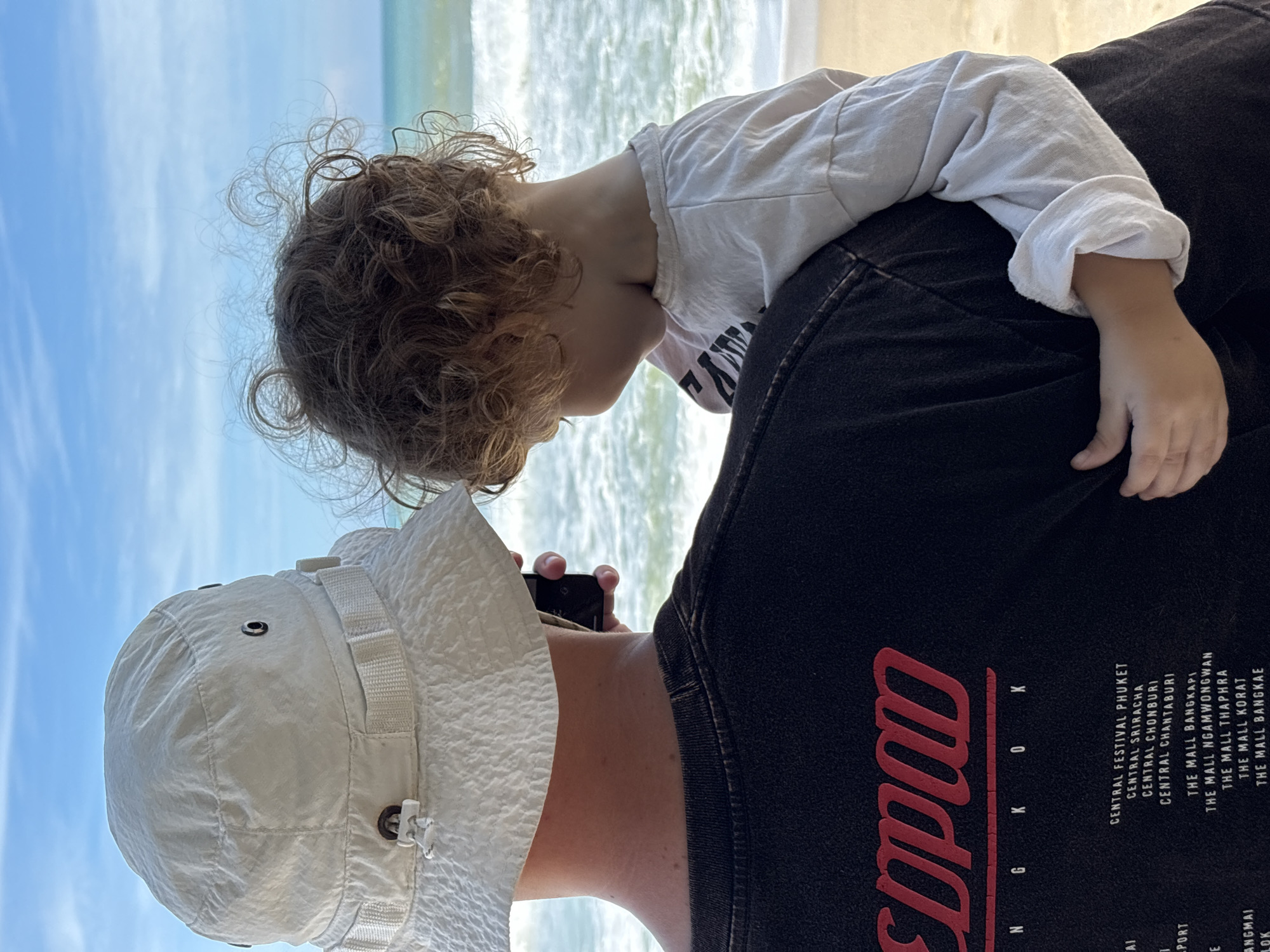Two individuals, an adult and a child, are positioned on a sandy beach, facing away from the viewer towards the ocean under a partly cloudy blue sky. The adult, wearing a white bucket hat and a black t-shirt with red text, is holding a mobile phone. The child, with curly brown hair, is being held by the adult, wearing a light-colored long-sleeved shirt. The ocean has moderate waves and the beach extends into the background. The t-shirt of the adult has text that appears to list locations, including "CENTRAL FESTIVAL PHUKET", "CENTRAL SRIRACHA", "CENTRAL CHONBURI", "CENTRAL CHANTABURI", "THE MALL BANGKAPI", "THE MALL NGAMWONGWAN", "THE MALL THAPHRA", "THE MALL KORAT", and "THE MALL BANGKAE". Based on the presence of "CENTRAL FESTIVAL PHUKET", a prominent shopping mall and tourist destination, the location is consistent with Choeng Thale, Phuket, Thailand.