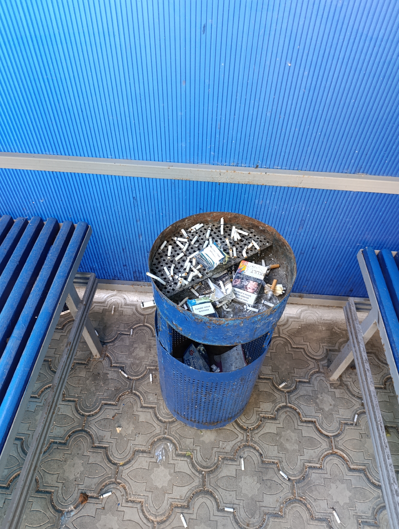 The image depicts an outdoor ashtray filled with numerous cigarette butts, located in the town council of Dubăsari, Moldova. The ashtray is a blue, perforated metal bin, showing signs of rust and wear. It is overflowing with cigarette butts, some still smoldering, and discarded cigarette packs. The ashtray sits on a patterned paved surface. To the left of the ashtray, a blue, slatted bench is visible. Overhead, a blue, corrugated roof structure is present, supported by a metal beam. The overall scene suggests a public outdoor smoking area that is frequently used.