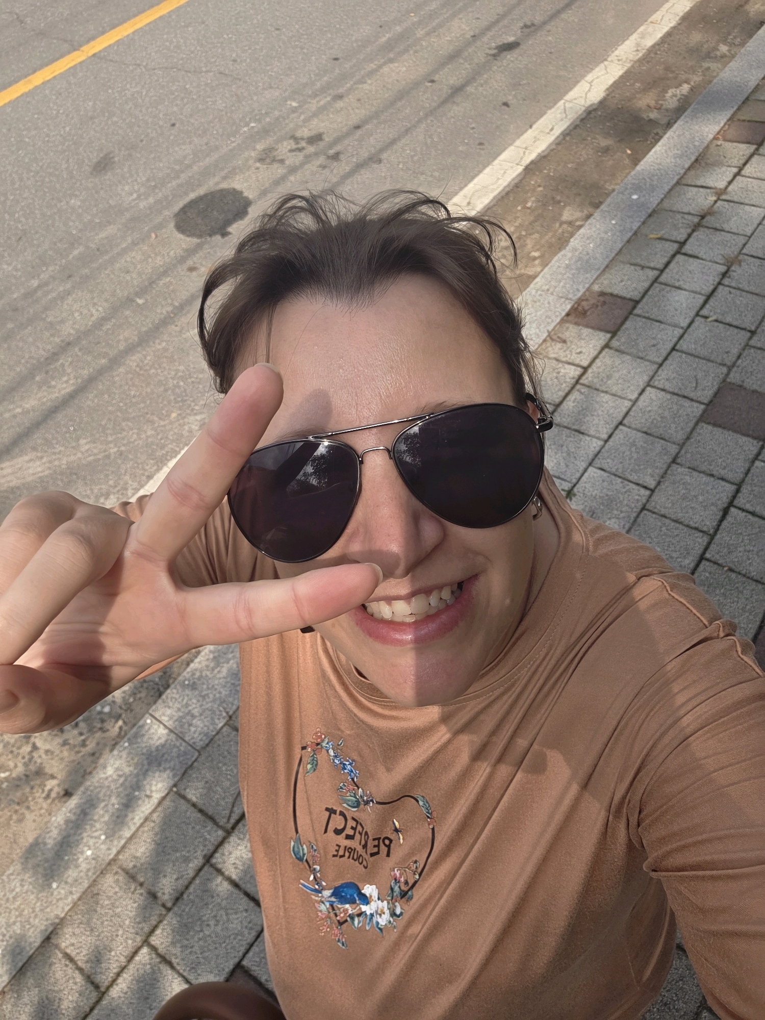 The image shows a woman, viewed from a high angle, standing outdoors on a paved surface. She is wearing sunglasses and a light brown long-sleeved shirt with a floral design and the text "PERFECT COUPLE" printed on it. The woman is smiling and making a peace sign with her left hand. The background consists of asphalt and a grey paved walkway, suggesting an urban or suburban environment. A yellow line is visible on the asphalt. The location is identified as Dunnae-myeon, South Korea, an area known for its natural scenery, but the image itself does not provide specific geographical markers to confirm this. The lighting suggests it is daytime, with shadows cast on the ground.