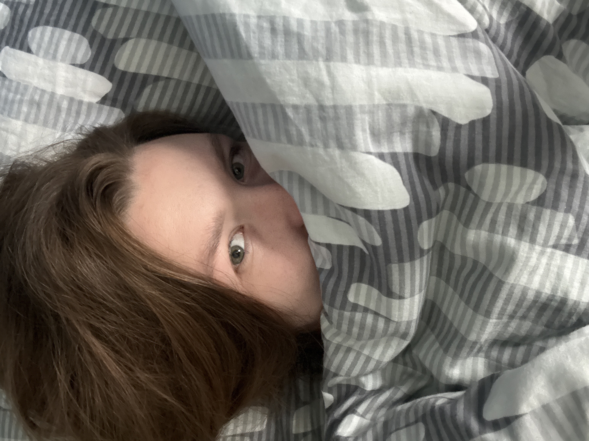 A person's face is partially obscured by a comforter with a striped and oval pattern in shades of gray and white. The visible portion of the face shows one eye with a green iris, and auburn hair is spread across the pillow. The image was taken indoors, likely in a bedroom in Izhevsk, Russia.