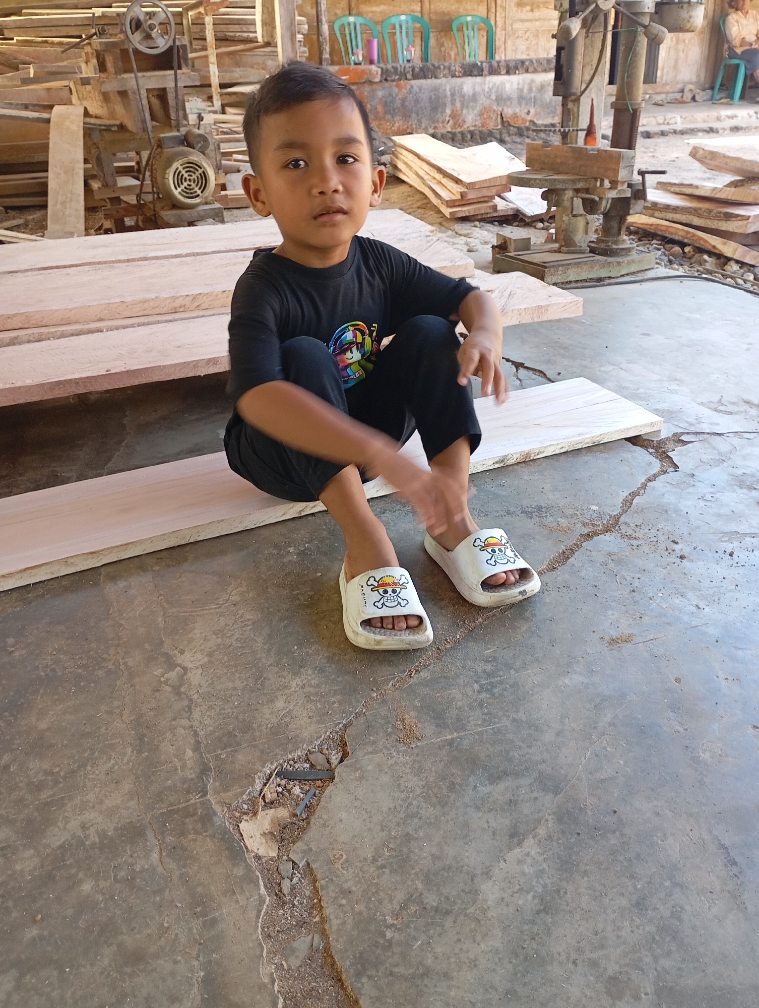 A young Indonesian boy sits cross-legged on a piece of unfinished lumber in what appears to be a woodworking shop in Ketepung, Indonesia. He is wearing a black t-shirt and black pants, with white sandals adorned with a skull and crossbones design. In the background, stacks of lumber and woodworking machinery are visible, suggesting the setting is an active workshop. The floor is concrete and cracked, with sawdust and debris scattered around. The overall lighting indicates daylight.