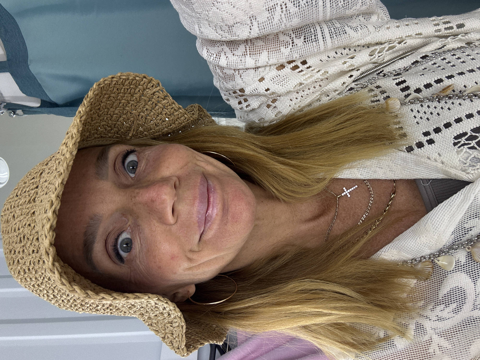 A woman is depicted, wearing a woven straw hat, gold hoop earrings, and multiple gold necklaces, including one with a cross pendant. She is also wearing a white lace garment. The background is a combination of white and blue. There is no indication of location.