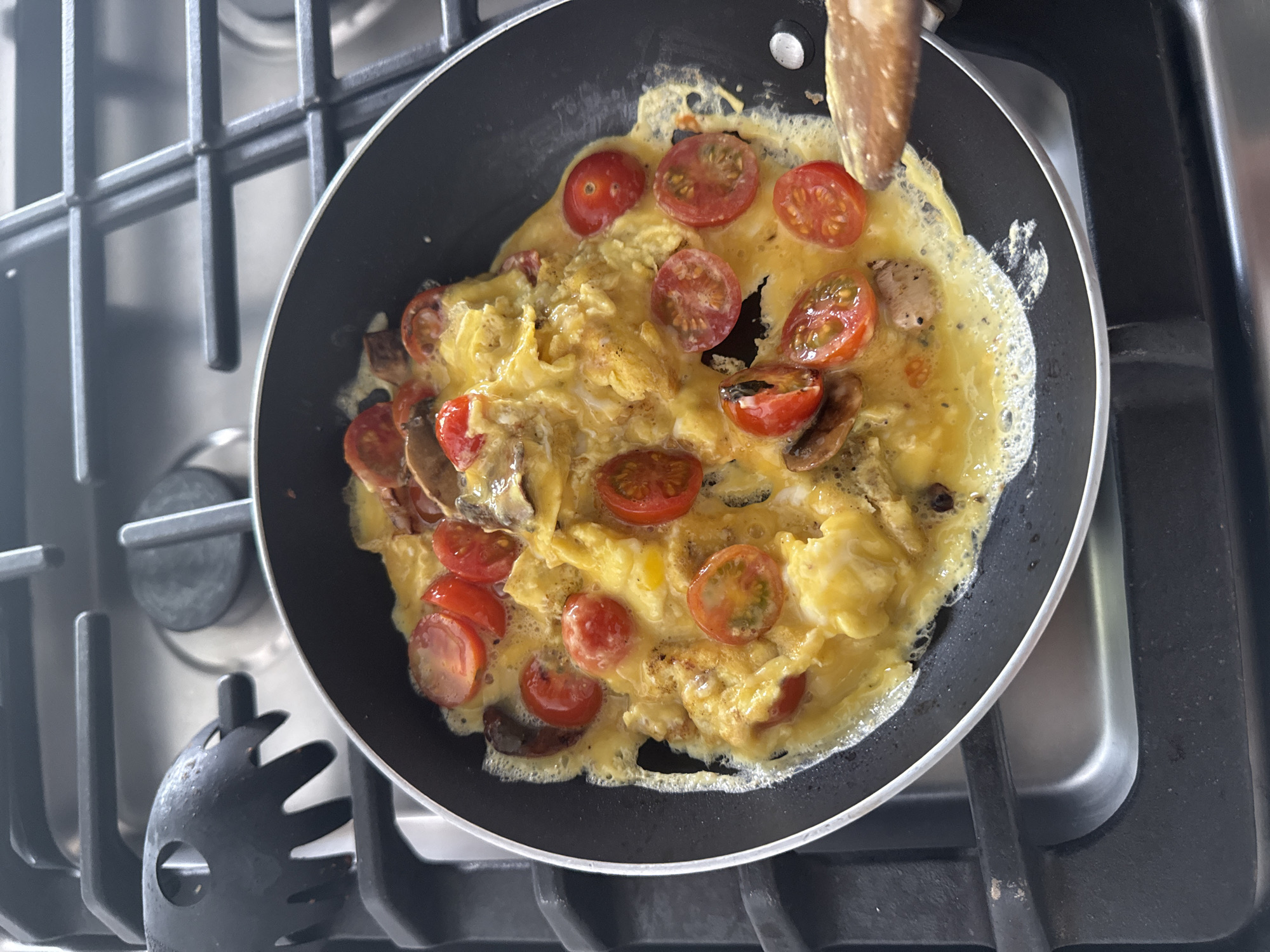 A top-down, close-up view captures a black non-stick frying pan on a gas stovetop, filled with food being cooked. The pan contains a vibrant dish of scrambled eggs or an omelet, appearing yellow and semi-solidified, with some frothy, liquid areas still visible. Mixed into the eggs are numerous bright red, halved cherry tomatoes and darker brown, sliced mushrooms. A wooden spatula or spoon is visible in the upper right of the pan, actively engaged in stirring or flipping the contents.
The frying pan has a black exterior and a thin, light-colored rim. It sits on the sturdy black grates of a gas range, which are prominent in the foreground and background. Beneath the grates, a reflective silver-grey stovetop surface is visible. In the lower-left corner, a distinctive black kitchen utensil with prongs, possibly a pasta server or draining spoon, rests on the grates. The scene conveys a moment of breakfast or brunch preparation in a domestic kitchen setting. No text is visible in the image.