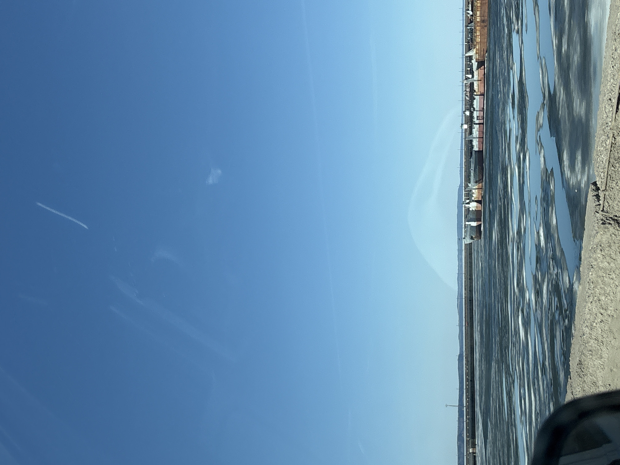 The image, correctly oriented, presents a wide landscape view under a clear, bright blue sky, characteristic of Chaikovsky, Russia. The sky dominates the upper portion of the frame, displaying a gradient from a deeper blue at the top to a paler shade closer to the horizon. A prominent white airplane contrail stretches across the upper left, with a few faint, wispy clouds scattered throughout.
The lower third of the image features a vast body of water, likely a river or lake, partially covered by broken and melting ice. The ice creates intricate patterns and reflections on the surface, indicating movement or thaw. Along the distant horizon, a low-lying landmass is visible, punctuated by a series of industrial structures. These include numerous cargo containers and various buildings, suggesting a busy port or industrial complex lining the waterway.
In the immediate foreground, a rough, textured patch of ground, possibly a gravelly shoreline or roadside, occupies the bottom right corner. A small part of a vehicle's interior, such as a side mirror or dashboard, is visible in the very bottom right, indicating the photo was taken from inside a car.
The scene captures a bright, sunny day, likely midday, with the sunlight strongly reflecting off the icy water. The presence of ice suggests cold weather, perhaps late winter or early spring. No people are visible, and no specific activity or event is occurring. There is no visible text in the image.
