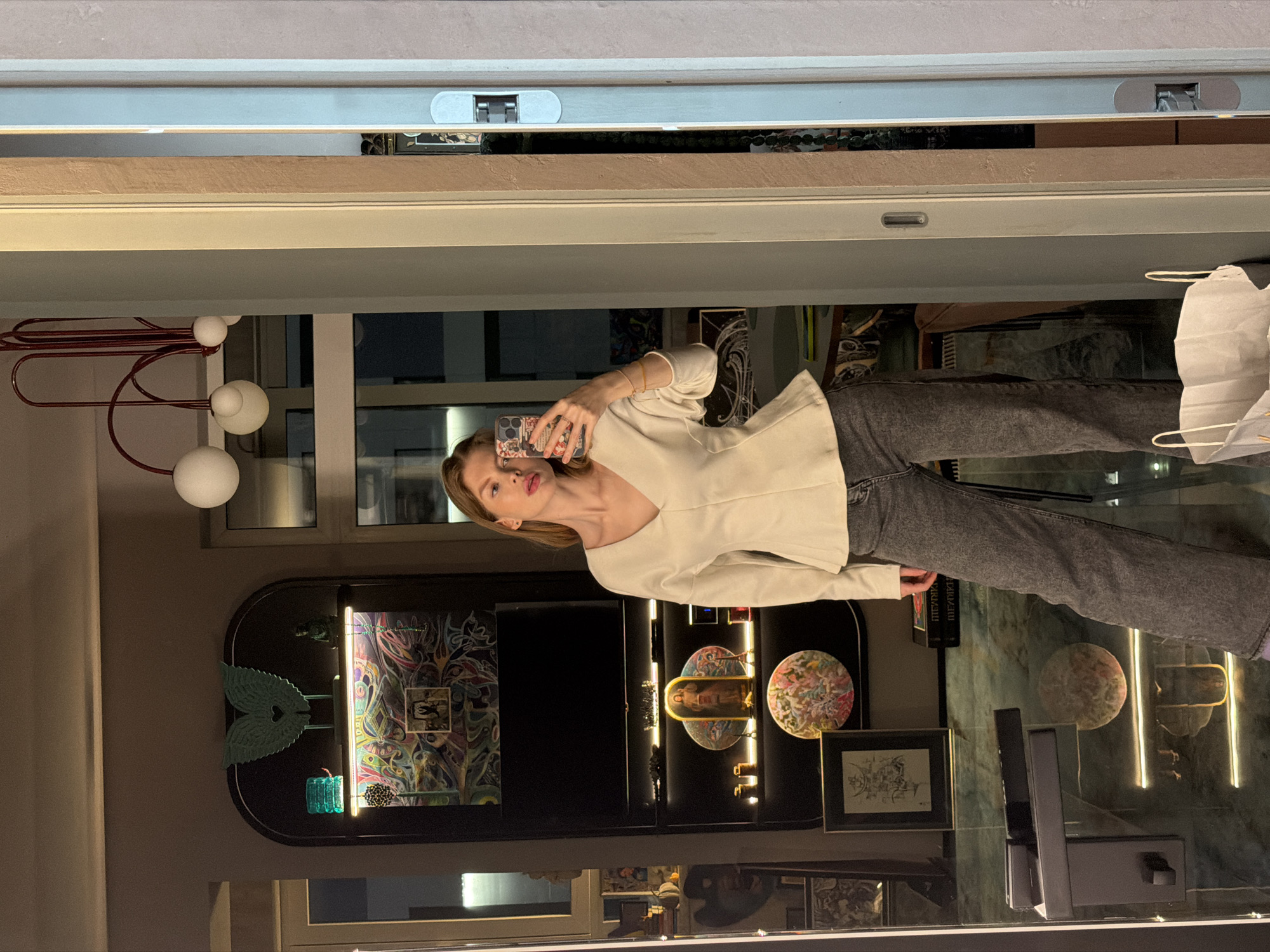 A woman is taking a selfie in a mirror inside a building. The walls and floors are stone-like. A tall shelving unit is visible, featuring artwork and decorative objects, some illuminated. A red-and-white lamp fixture stands on the left. Through the window are visible lights that could be the city lights of Moscow, Russia. The woman is wearing a cream-colored top and gray jeans.