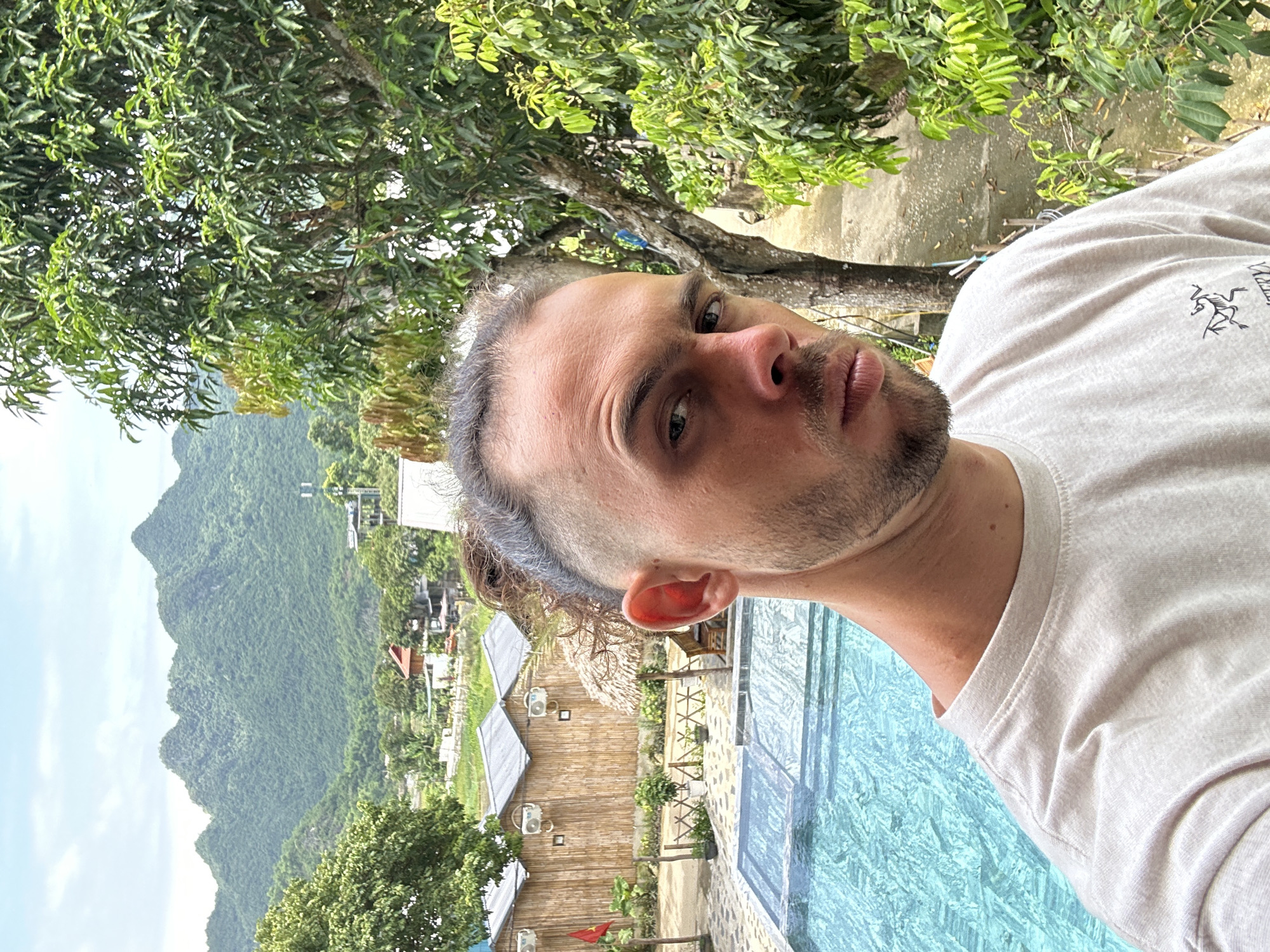 A man with his hair pulled back in a bun and a short beard takes a selfie in front of a scenic backdrop of lush green mountains and trees. He is wearing a light-colored t-shirt with a logo on the chest. To his left, a swimming pool with turquoise water is visible, along with a few thatched-roof huts and some wooden fencing. The scene appears to be a resort or guesthouse located in Xóm Khán, Vietnam, characterized by its natural beauty and tranquil atmosphere. The sky is partly cloudy, suggesting a bright but potentially humid day. The man's gaze is directed at the camera, with a neutral expression.