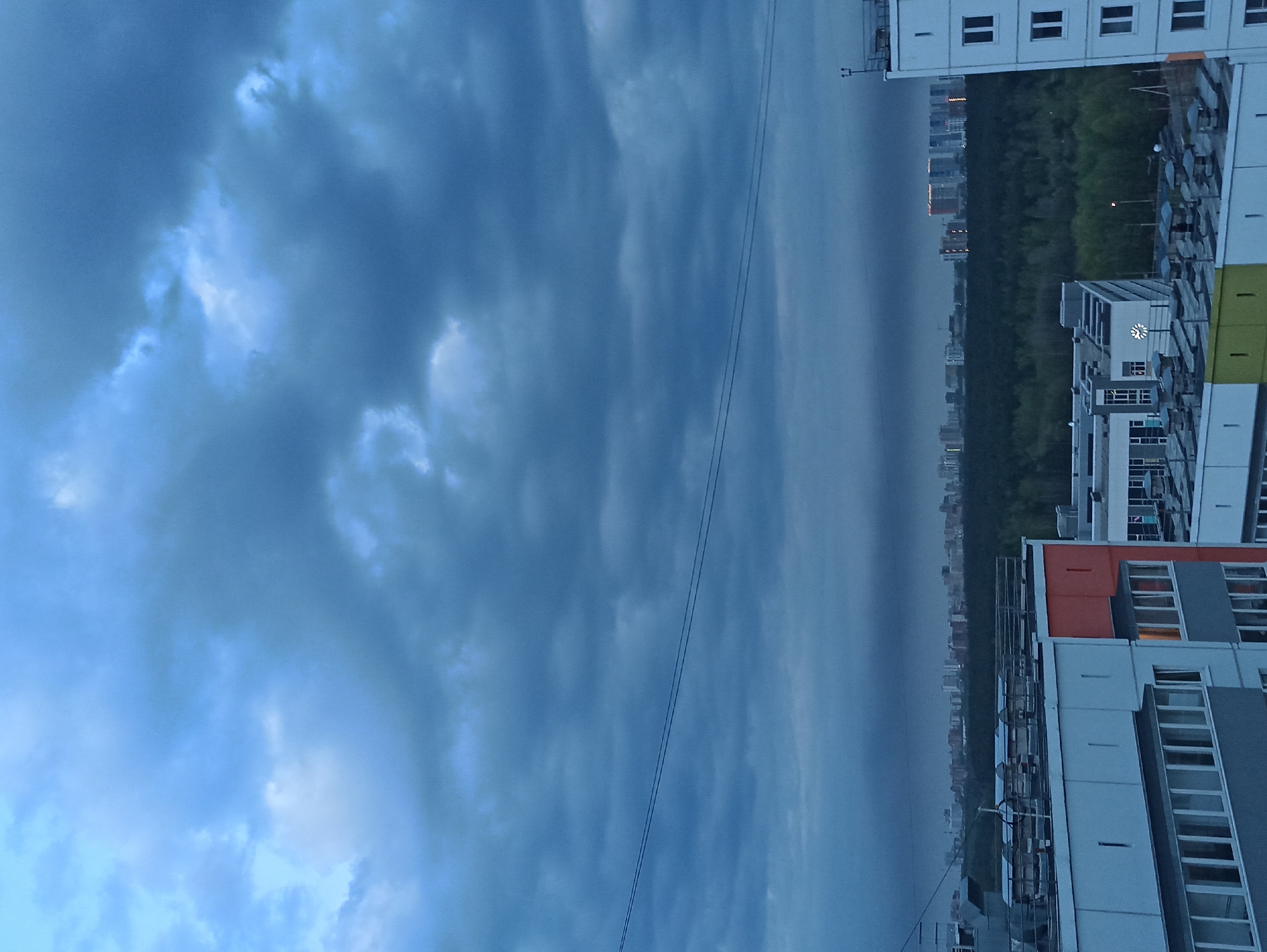 The image shows a view from a high vantage point in Кондратово, Russia, looking across an urban landscape towards a cloudy sky. A multi-story residential building occupies the lower portion of the frame, exhibiting a modern architectural style with a facade composed of white and grey panels, punctuated by windows and occasional orange accents. Behind the building, a dense green forest or park stretches towards the horizon. Beyond the trees, the skyline of a distant city is faintly visible, marked by a series of high-rise buildings. The sky takes up the majority of the frame, dominated by dramatic, dark storm clouds. Overhead power lines stretch across the view toward the building.