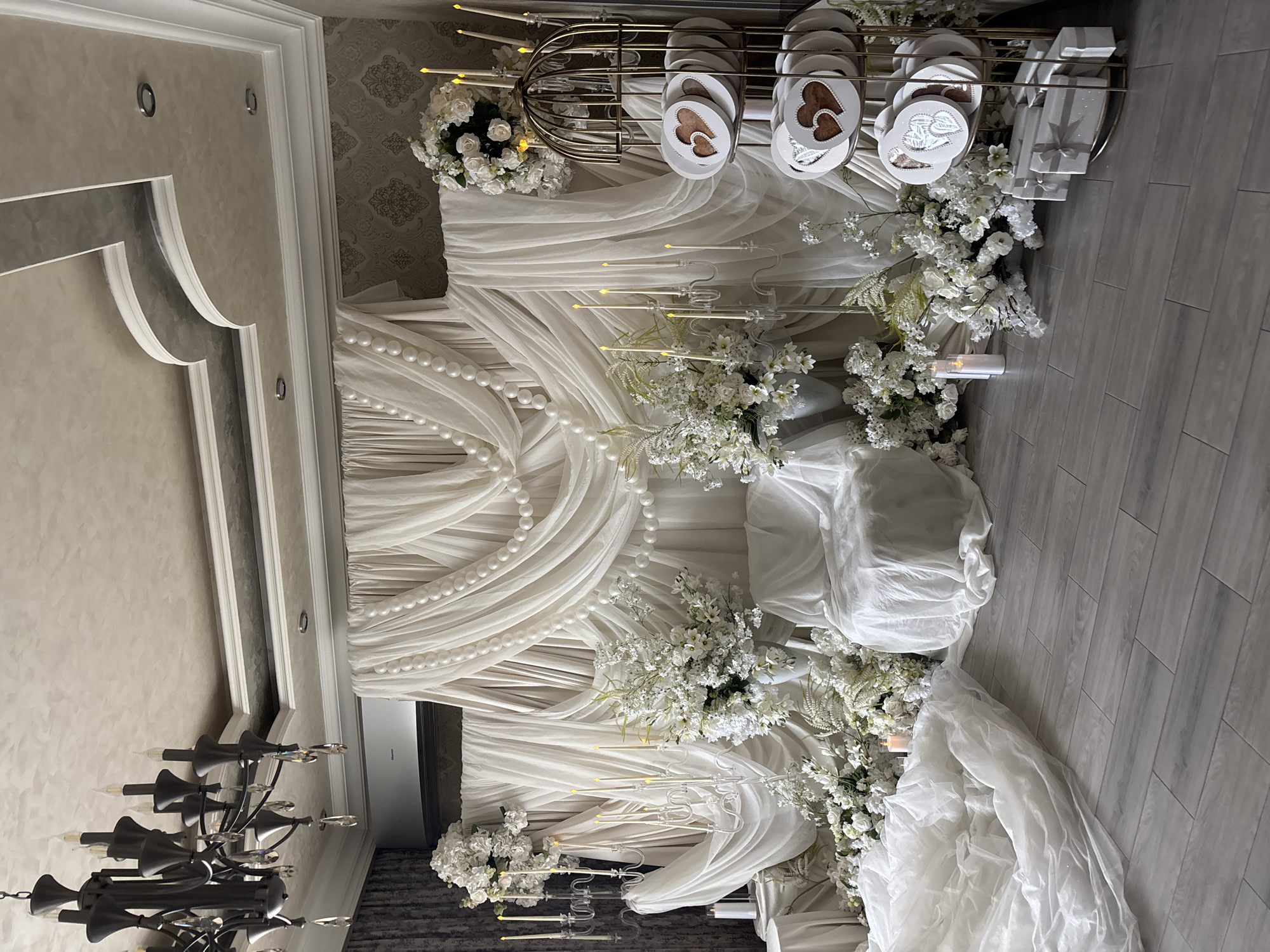 The image shows an elaborate wedding or event decoration, likely indoors, in what seems to be a reception hall. The dominant color theme is white.
The backdrop is composed of draped white fabric, cascading down in layers and accented with strings of pearls. White floral arrangements are interspersed throughout, adding to the elegant and romantic atmosphere. A golden birdcage structure sits atop the backdrop. Within the cage and on the table are objects that appear to be wedding favors, including what look like small heart-shaped gifts or cards.
There are white candles arranged on ornate candle holders.
The room's floor is light-colored and appears to be made of wood panels. A black chandelier is visible in the foreground, hanging from the ceiling.
Given that I don't have specific information to confirm the location, the information about Yerevan, Armenia, is not verifiable based on the visual content alone.