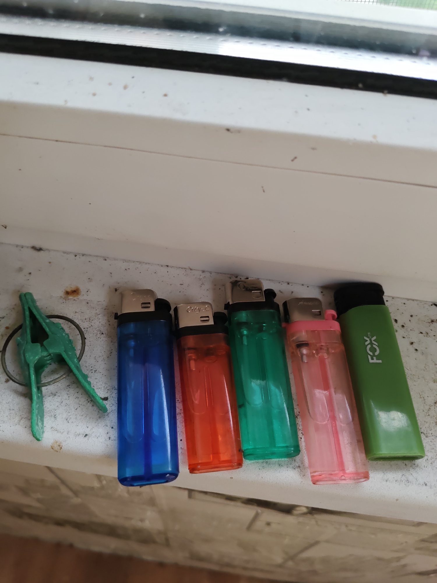 A group of six lighters and a green clothespin are arranged on a white windowsill. The lighters are, from left to right: blue, red, green, pink, and green with a black cap that reads "FOX". The clothespin is to the left of the lighters. The windowsill is dusty and shows some signs of wear. The background is a blurry view of a window. The scene appears to be indoors. This image was taken in Rostov-on-Don, Russia.