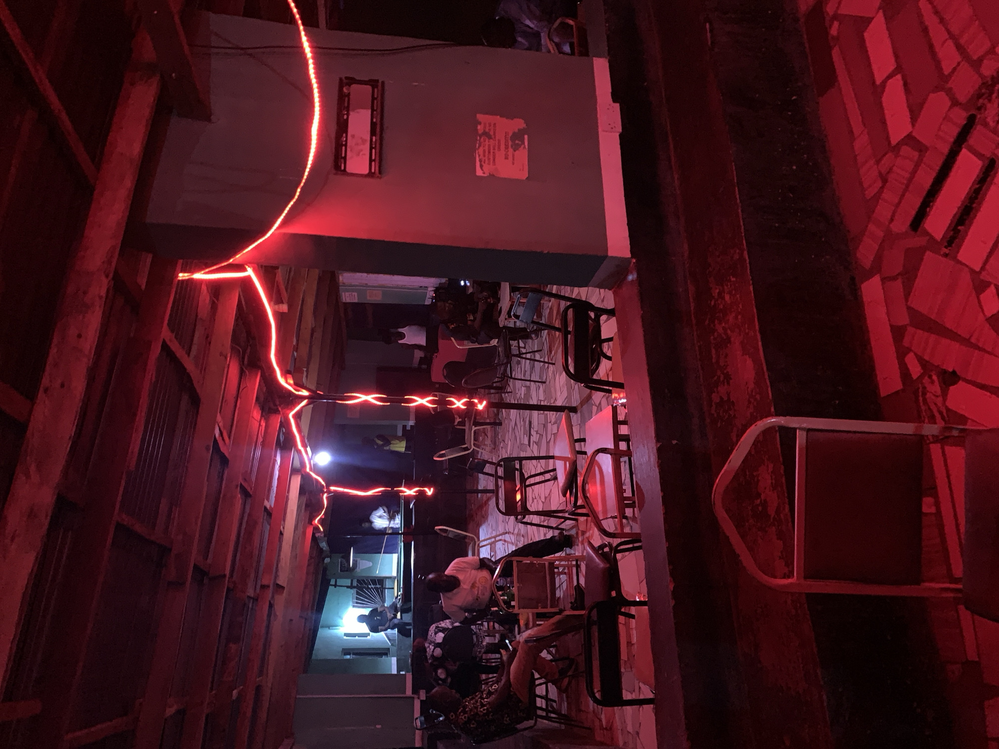View of a dimly lit outdoor bar/eating area. Several people are seated at tables and chairs. Red neon light fixtures illuminate the scene. Wood panels on the left and red tiles are visible on the right. The scene is in New Stadium, Nigeria.