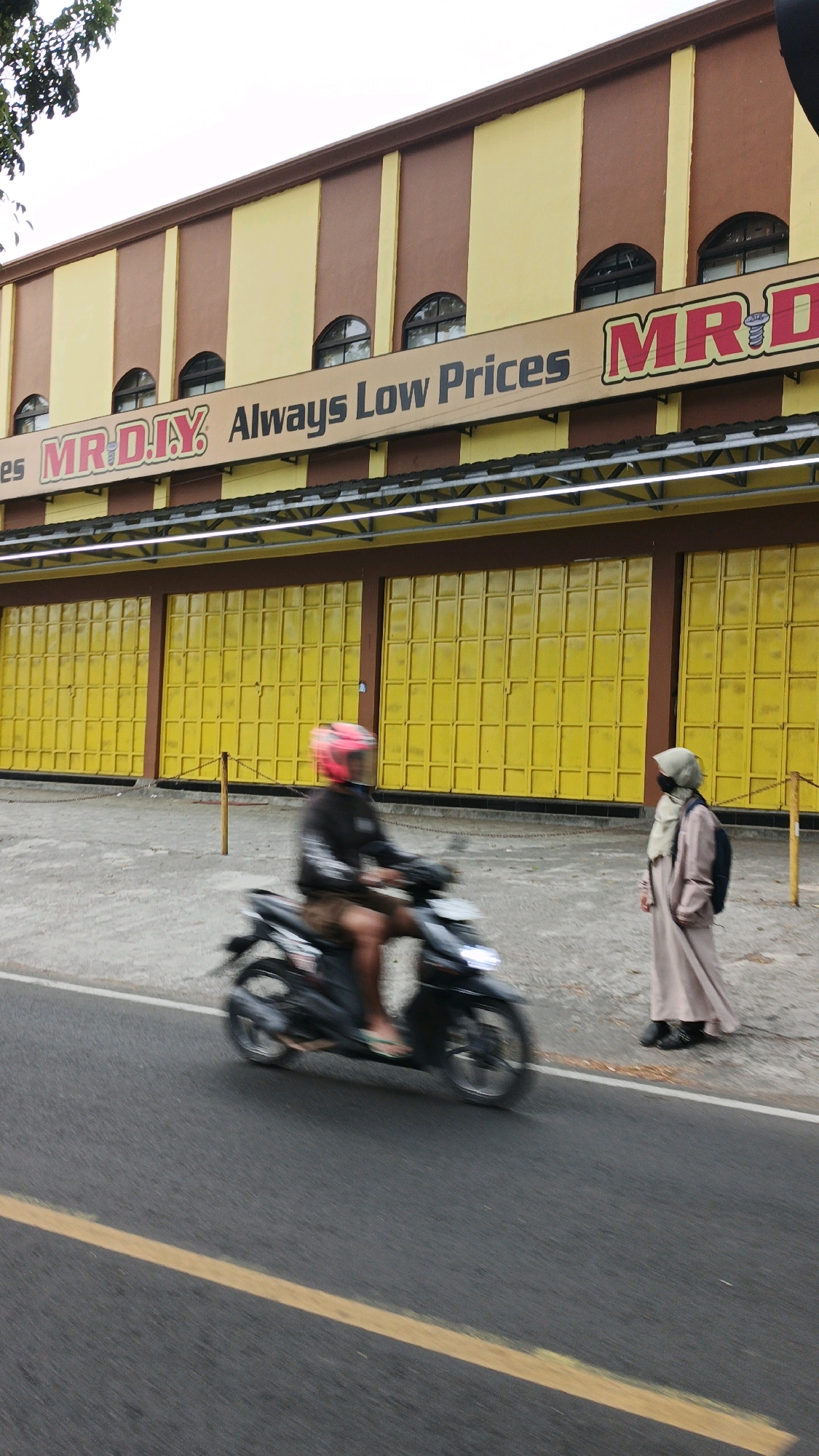 The image shows the MR. D.I.Y. store in Desa Wonoharjo, Indonesia. The building has a brown and yellow striped facade, with a banner that reads "MR. D.I.Y. Always Low Prices". The store's entrance is covered by large yellow metal shutters. In the foreground, a person is riding a motorcycle past the store. The motorcycle is moving, causing motion blur. To the right of the motorcycle, a person wearing a hijab and a long robe stands on the sidewalk, facing the store. The road in front of the store has a yellow dividing line. Trees are visible on the upper left side of the image.