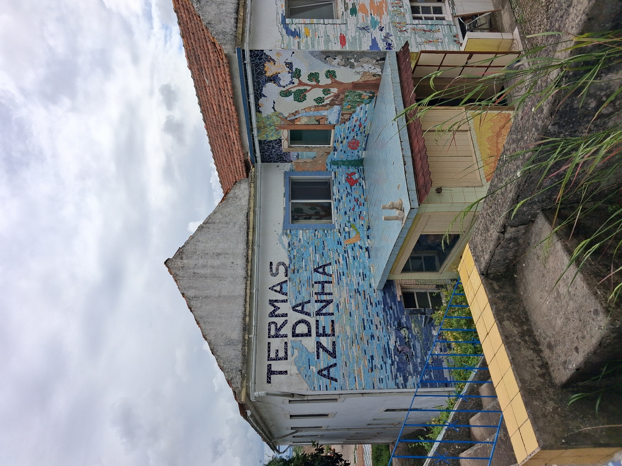 The image shows a building in Soure, Portugal, identified by a vertical mosaic that reads "TERMAS DA AZENHA". The building has a light-colored facade with tiled mosaics depicting different scenes, including a tree and water motif, and colorful fish and dragonflies. The building has windows with light blue trim. A red-tiled roof is visible. Adjacent to the building, there's a concrete wall with grass growing. Blue metal railings and steps are in front of the building. The sky above is overcast with many clouds.