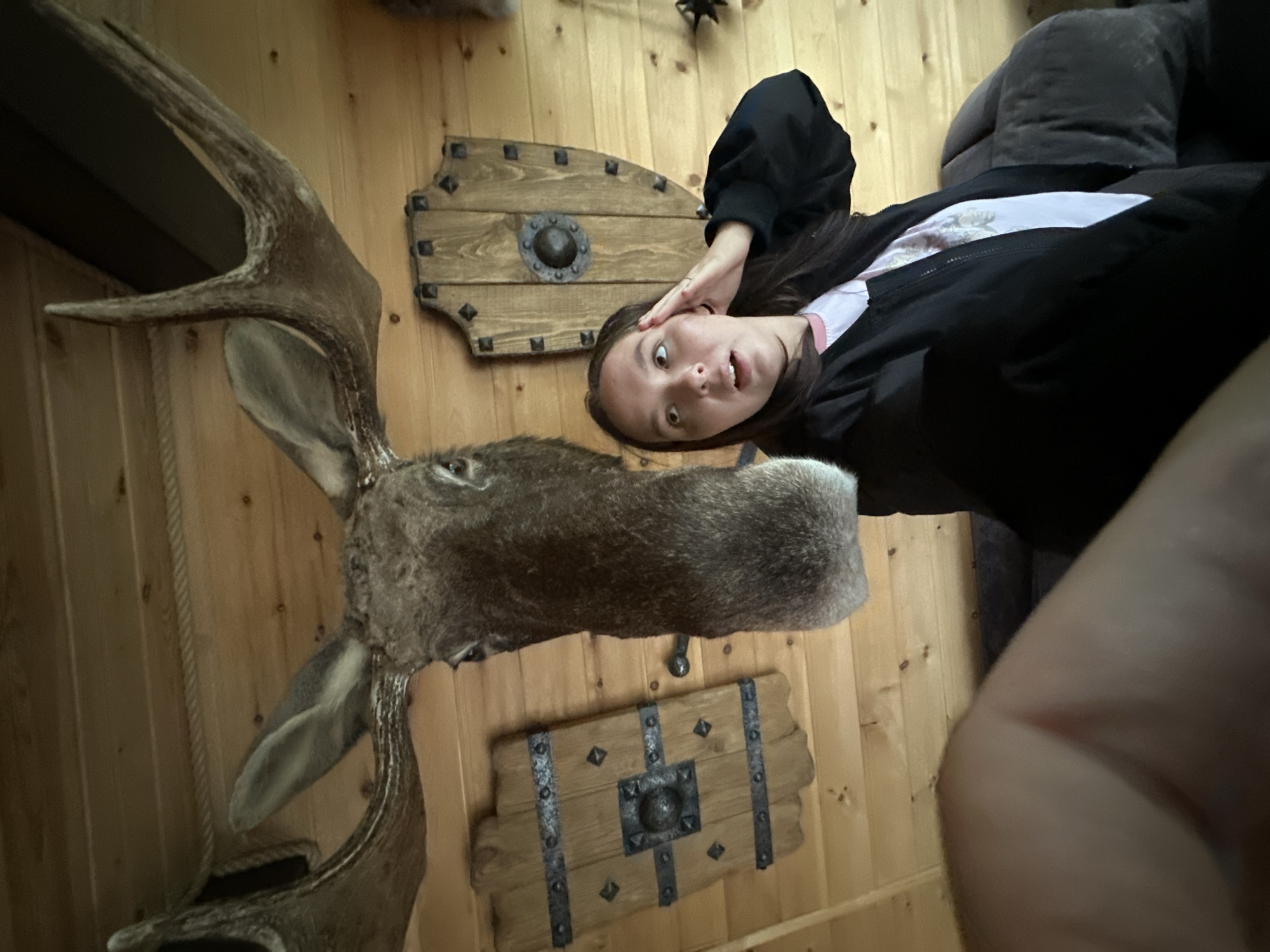 A young woman with dark hair and a light-colored t-shirt and black jacket is posing for a selfie in a room with wood-paneled walls. She has her right hand on her forehead and her mouth is slightly open in a surprised expression. Behind her and to her left, a mounted moose head with large antlers is prominently displayed. To her left and right are decorative wooden shields with metal accents. The room appears to be dimly lit, suggesting an indoor setting. There is no indication of the city of Karsha, Russia within the image.