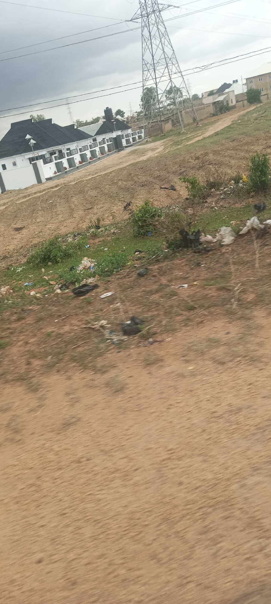 The image captures a scene on the outskirts of Jos, Nigeria, marked by a juxtaposition of residential development and environmental degradation. Several newly built, white-walled houses with black roofs line the left side of the frame, partially obscured by an electricity pylon in the background.
The foreground reveals an unpaved, sandy area with sparse vegetation mixed with scattered litter and plastic debris. The lower portion of the image is blurred, suggesting motion, possibly captured from a moving vehicle. The overcast sky provides a dull, diffused light, creating a somewhat bleak atmosphere.