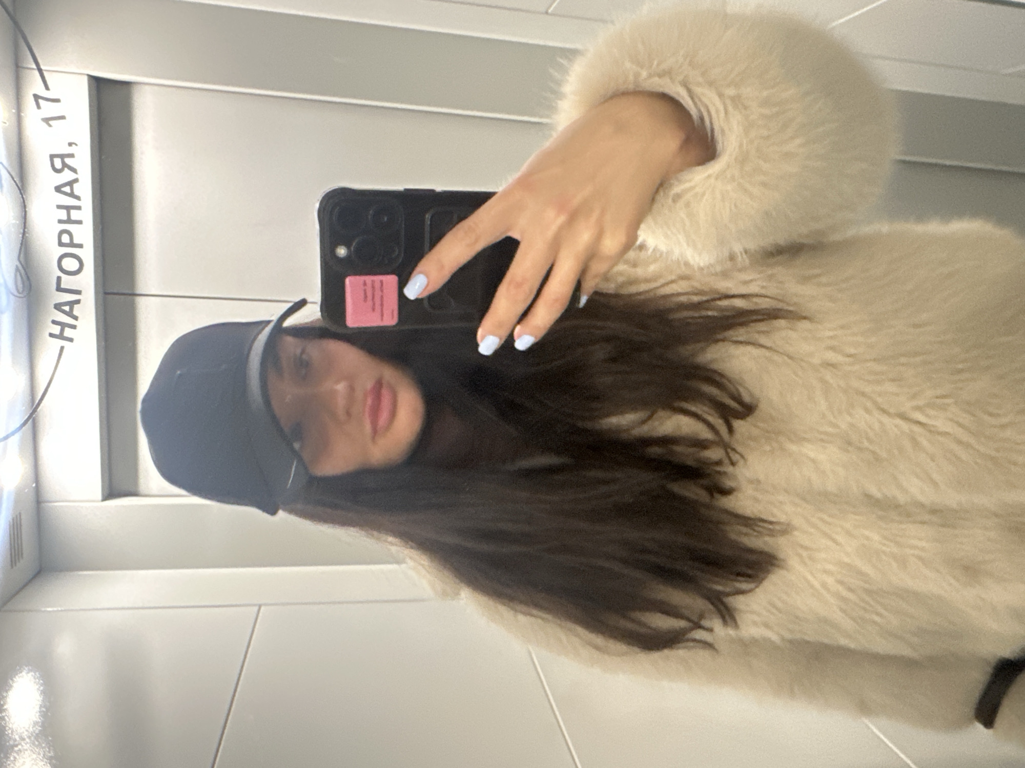 A woman is captured in a mirror selfie, positioned vertically in the frame. She has long, dark hair and is wearing a light, fluffy, possibly faux fur coat and a dark baseball cap. Her face is partially visible, showing light-colored lips. She holds a dark-colored smartphone, appearing to be an iPhone Pro model, with her left hand, which has light blue nail polish. A small, rectangular pink sticker is affixed to the back of the phone case.
The background, reflected in the mirror, consists of light grey or white panels with subtle horizontal lines, suggesting an indoor setting like an elevator or a modern hallway. The woman is looking into the phone's camera within the reflection, capturing her image.
Prominently visible on the mirror's surface, to the left of the woman's reflection, is text in Cyrillic letters: "НАГОРНАЯ, 17". This translates to "NAGORNAYA, 17," indicating a street name and building number. Given the context of Khimki, Russia, this likely refers to an address in that vicinity. The overall impression is a casual, contemporary portrait taken in an urban indoor environment.