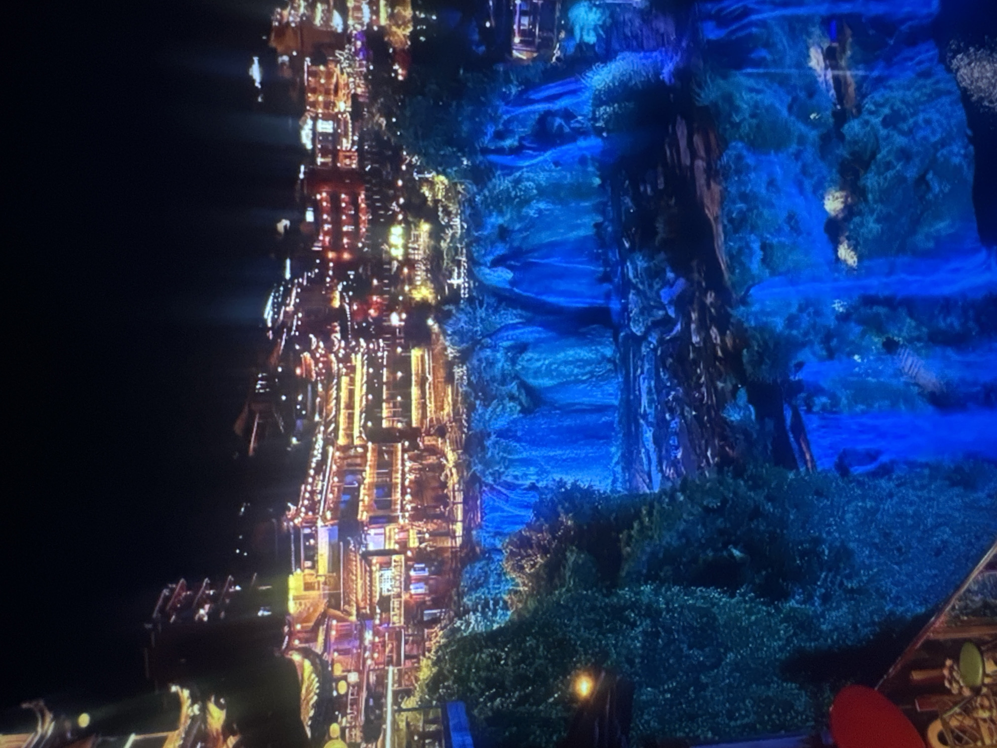 This is a nighttime, medium-shot image of a city built on a hillside overlooking a brightly lit waterfall. The city is illuminated with numerous warm-toned lights, creating a festive atmosphere. Traditional-style architecture with multiple levels and intricate details is visible, with many windows and balconies adorned with lanterns or string lights. The waterfall, illuminated with vibrant blue light, cascades down multiple tiers, surrounded by lush greenery. The water reflects the blue light, creating a mystical and dramatic effect. In the foreground, there appears to be a balcony or viewing area with some furniture, including a red heart-shaped object, a stool, and a railing. The sky is dark, indicating it is nighttime.