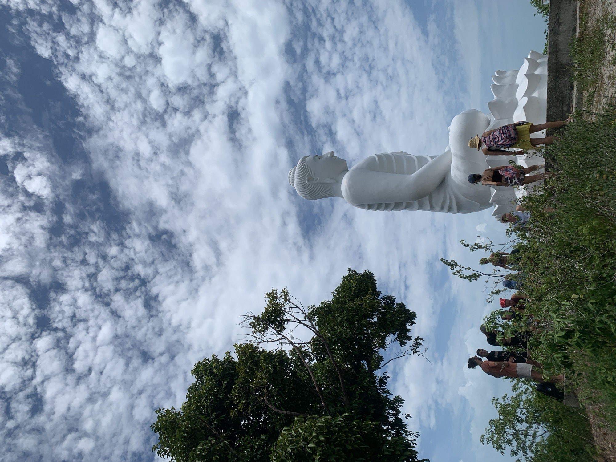 In a wide shot, a colossal white Buddha statue sits serenely in profile against a bright, cloudy sky. The statue, positioned on a hillside, is partially obscured by lush green foliage and several people gathered in the foreground. The people, dressed in casual attire, are observing the statue and the landscape. The environment appears to be a natural setting with a hint of man-made structures, possibly a temple or monument site. The image conveys a sense of scale and wonder, with the immense statue dwarfing the human figures and the surrounding vegetation. The location is stated to be unknown, Thailand.