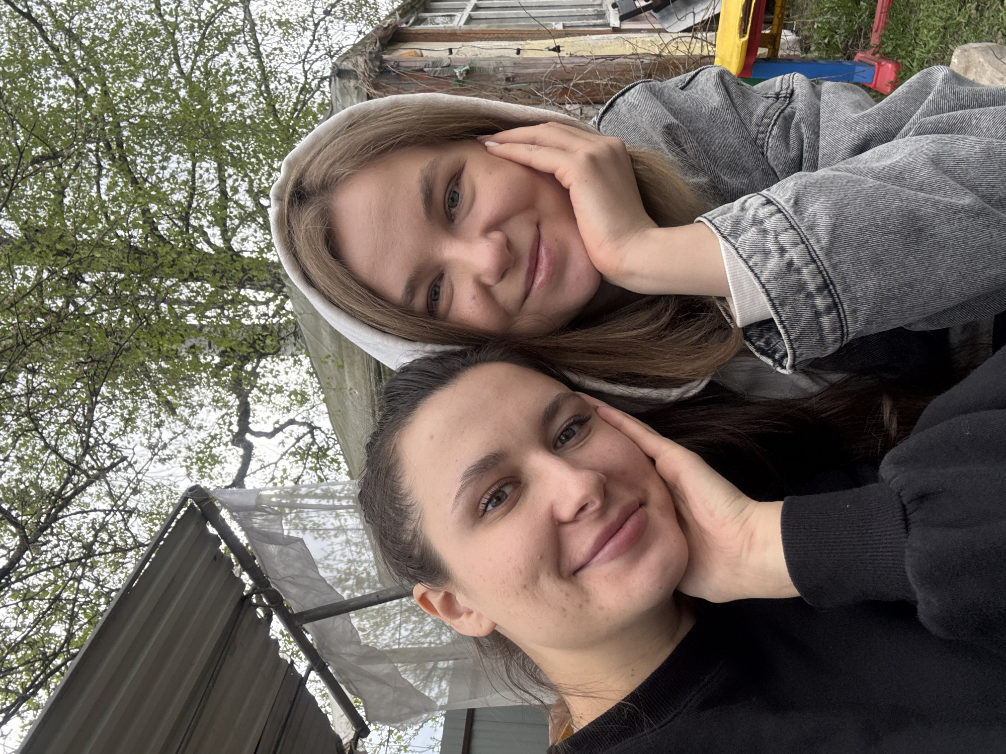Two fair-skinned women are posing closely together for a selfie outdoors. The woman on top is wearing a light gray hooded jacket and has her hand on her cheek, smiling. The woman below, wearing a black sweater, is also smiling and similarly has her hand resting on her cheek. The background includes trees with green foliage, a section of a building, and what appears to be part of a child's playset in the blurred distance.
