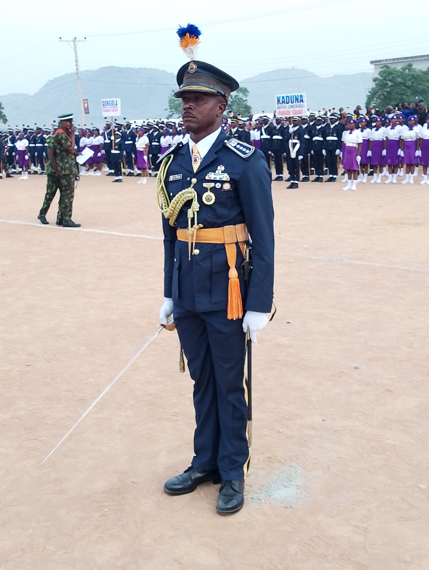 The image depicts a uniformed officer, presumably in Kubwa, Nigeria, given the banners in the background, standing in a parade formation on a dusty field. The officer wears a navy blue uniform, peaked cap with a plume, light-colored tie, gold-colored belt with a tassel, white gloves, and black shoes. He is holding a sword. In the background, there are rows of uniformed personnel and civilians in white and purple uniforms. Banners reading "GONGOLA" and "KADUNA" are visible. A man in military camouflage stands to the left of the central figure. Mountains are faintly visible in the far background under an overcast sky.