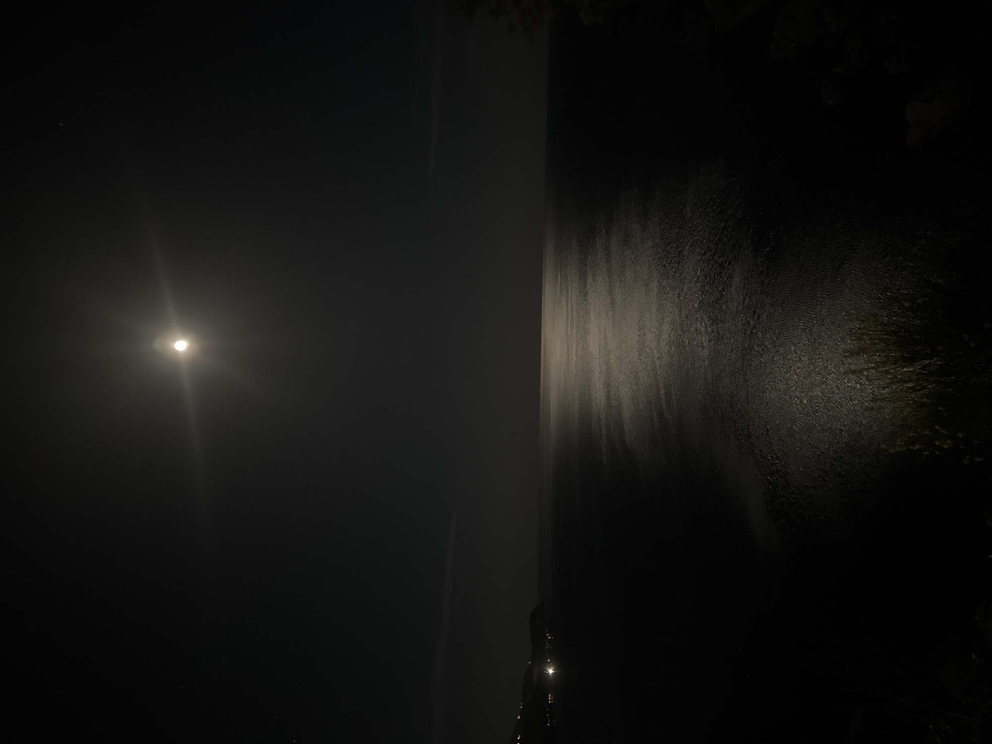 A night scene in Rafailovići, Montenegro, featuring a full moon over the Adriatic Sea. The moon is positioned in the upper left of the frame, casting a bright reflection on the dark water. The right side of the image shows the textured surface of the sea closer to the shore, with darker areas indicating shadows or deeper water. A few faint lights are visible along the shoreline. The sky is dark and mostly clear, with a few visible stars. The overall mood is serene and dark.