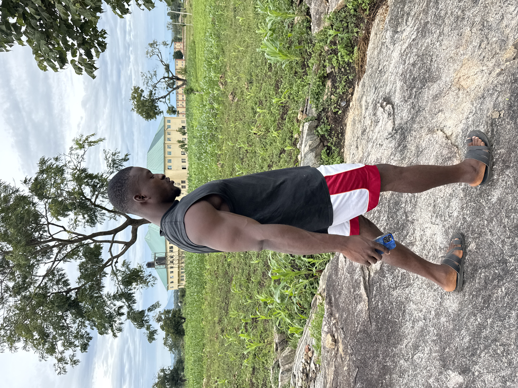 A man stands on a large rock in a grassy field. He is wearing a black sleeveless shirt and red and white shorts. He has dark skin and short black hair. In his right hand, he holds a blue mobile phone. To his left, a large tree with green leaves partially obscures the view of a yellow building in the background. The sky is overcast. The setting appears to be a rural area, likely in Akwanga, Nigeria. The man's posture suggests he is posing for a photo or looking out at the landscape. There is no visible text in the image.