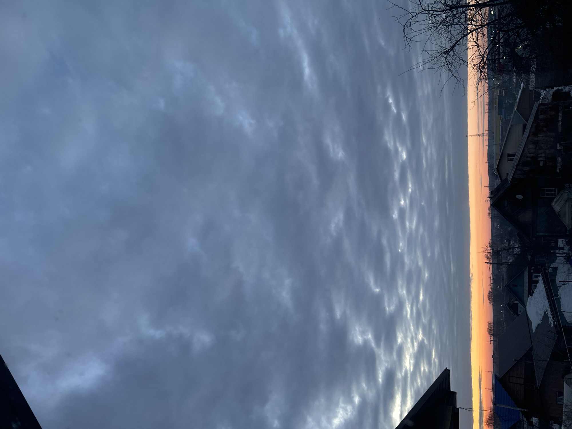 An elevated view captures an overcast dawn or dusk over a residential area in Chaikovsky, Russia. The upper two-thirds of the frame are dominated by a grey-blue sky filled with stratocumulus clouds. A distinct, linear band of orange and yellow light indicative of a low sun angle marks the horizon, gradually fading into the grey-blue clouds above. Below this horizon, the urban landscape consists of numerous single-story and two-story houses, primarily with dark pitched roofs, some retaining patches of snow. Visible roof colors include dark grey, blue, and brown. Bare deciduous trees are present on the right side of the frame and further in the distance. A tall, slender lattice transmission tower and other industrial structures are silhouetted against the bright horizon in the mid-ground. A dark roof overhang partially obscures the upper left corner of the image. No individuals are depicted. The scene suggests a cold weather season with snow present.