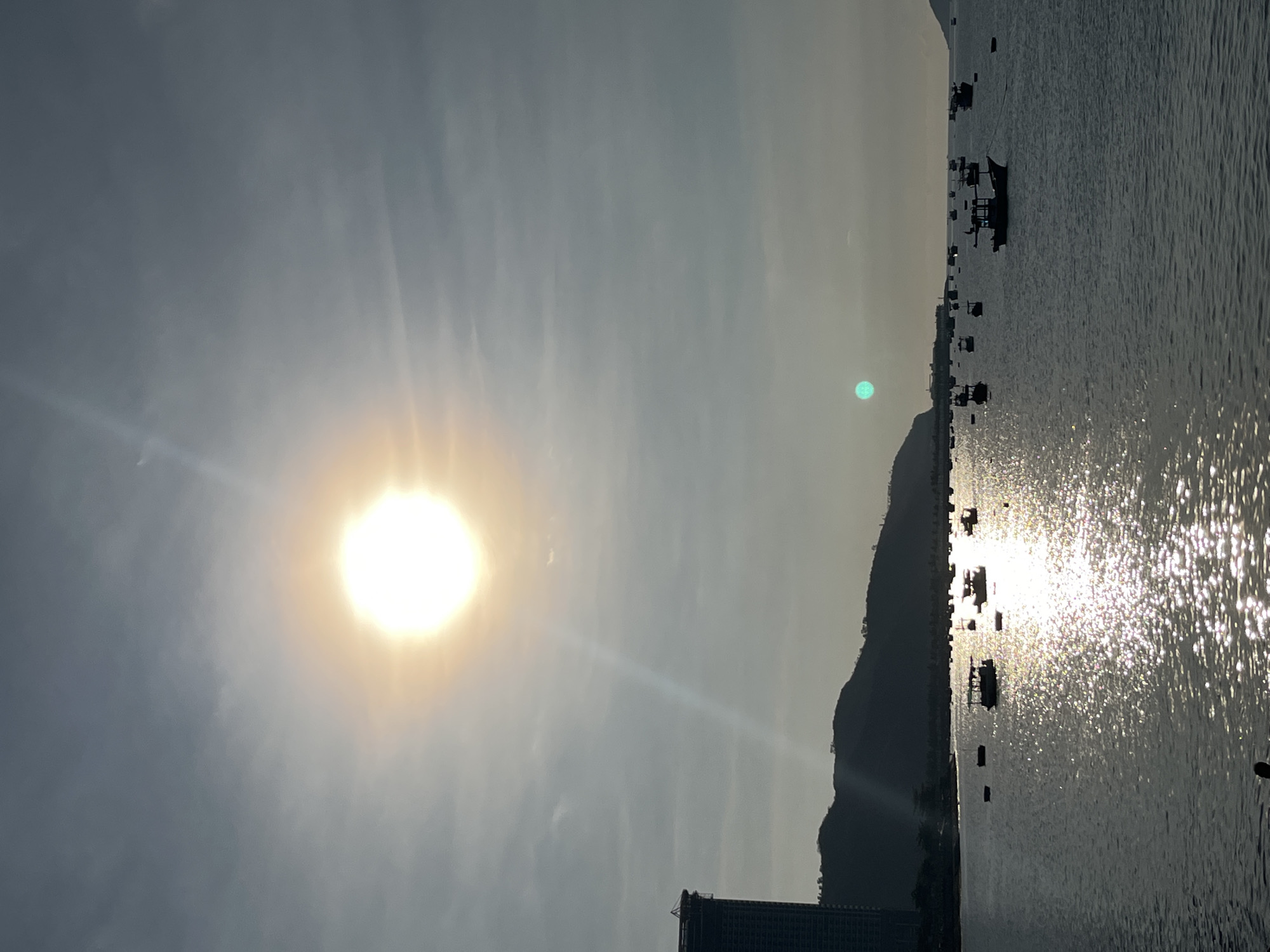 The image shows a sunlit seascape with various elements. The sun is bright and positioned prominently in the upper-left portion of the frame. The water reflects the sun's light, creating a shimmering effect. In the distance, there is a coastline or landmass with a prominent mountain silhouette. Small boats are scattered on the water. A building structure is visible at the bottom, partially obscured. There is a hint of Bac Nha Trang Ward, Vietnam.