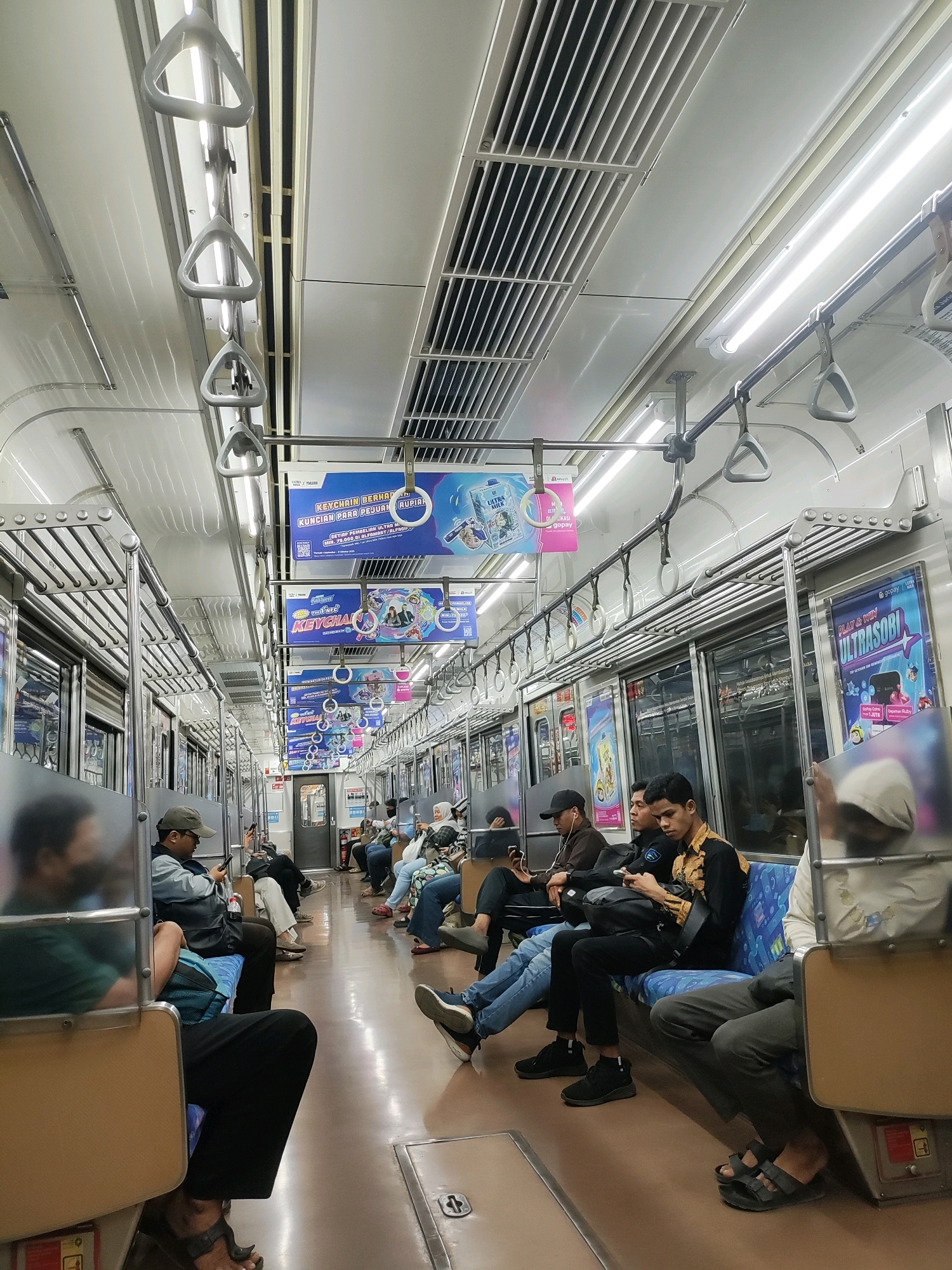 The image depicts the interior of a commuter train, likely in Mekarsari, Indonesia, during daytime. The train car is elongated with rows of seats on either side of a central aisle. Overhead, there are numerous white triangular handrails and fluorescent lighting fixtures. The walls and ceiling are predominantly white. Several advertisements are displayed on the walls above the windows.
In the foreground, passengers are seated on blue patterned seats. On the left, a person wearing a green shirt is seated with their back to the camera, partially obscured by a reflective surface. To their right, a person wearing a grey cap and a grey jacket is looking at their phone. Further down the aisle, other passengers are visible, some looking at their phones, others talking or resting.
In the midground, more passengers are seated. On the right side of the aisle, a person in a patterned shirt is looking at their phone, while another person in a black jacket is next to them. The flooring is a light brown, reflective material.
The overall atmosphere suggests a typical commute, with passengers engaged in individual activities or resting during their journey. The train car appears clean and well-lit.