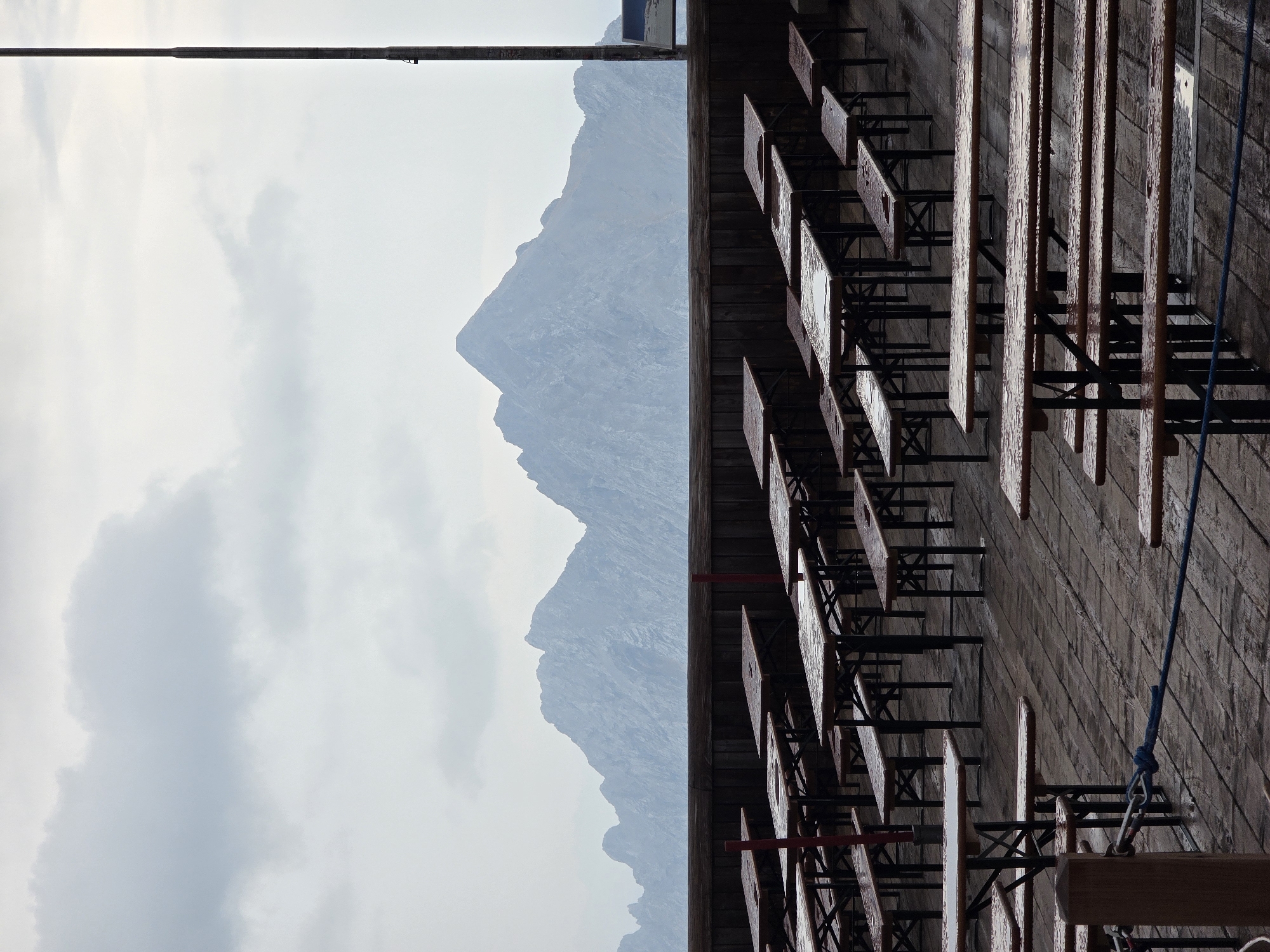 The image depicts an outdoor seating area with numerous empty wooden picnic tables and benches arranged on a wet wooden deck. The tables are made of wood with metal legs and appear to be covered in water droplets, suggesting it is raining or has recently rained. In the background, a dark wooden fence separates the seating area from a panoramic view of a mountain range under a cloudy, overcast sky. The mountains are rugged and appear to be covered in patches of snow. To the right of the frame, a tall, slender, dark-colored pole, possibly a light post or antenna, rises vertically into the sky. The overall atmosphere is somber and quiet due to the emptiness of the seating area and the gloomy weather. The location is identified as Garmisch-Partenkirchen, Germany.