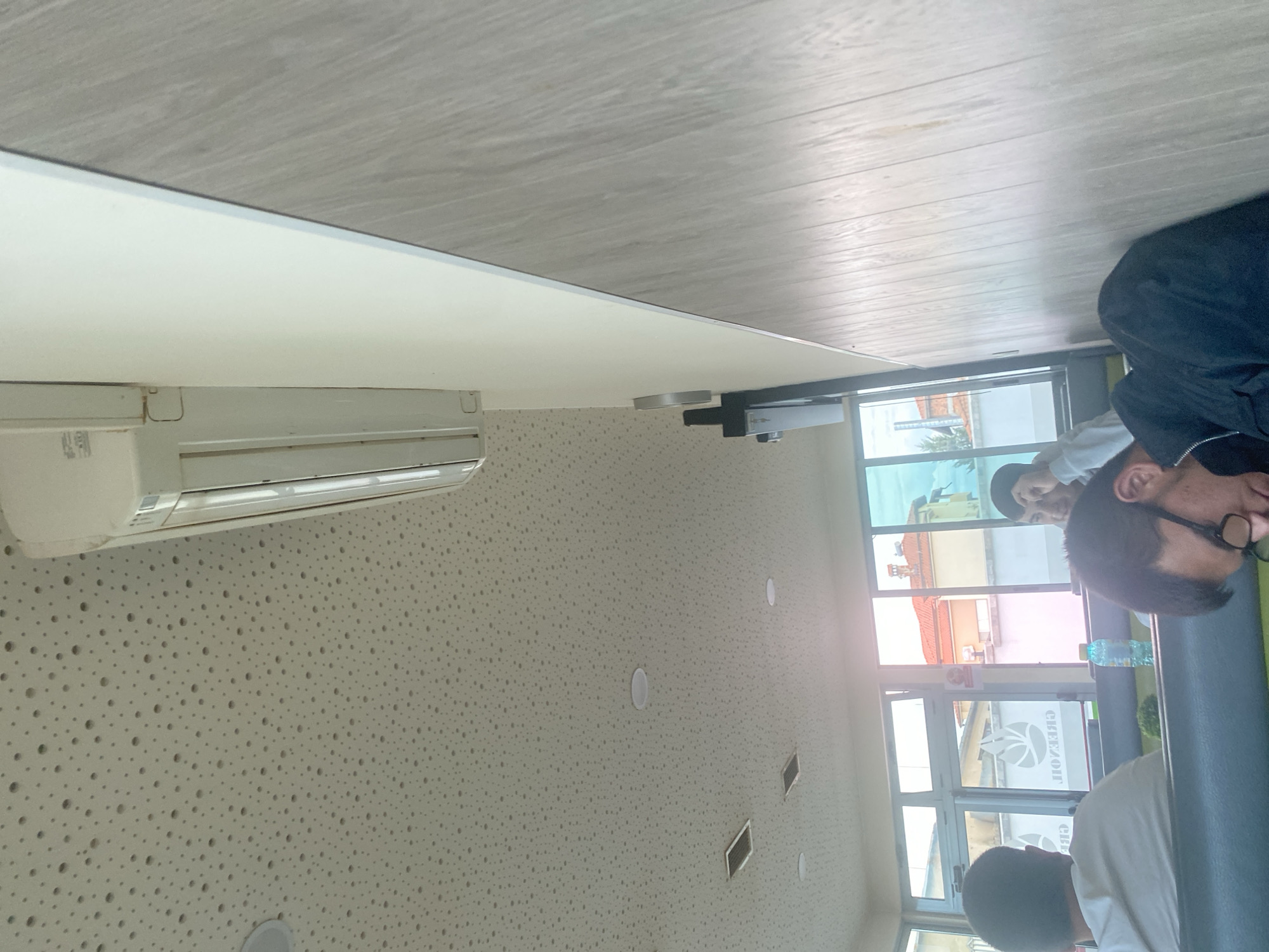 The image depicts the interior of a cafe in Ablanitsa, Bulgaria. The perspective is from a seated position, looking upwards and to the right. The ceiling is a textured white material with numerous small perforations. A white air conditioning unit is mounted high on the wall, extending horizontally. To the right, a wall with a wood-grain pattern runs vertically, with a light-colored trim separating it from the ceiling.
In the foreground and to the left, a person wearing a white t-shirt is seated, with their back to the camera. In the middle ground, through a large window, the exterior of buildings with orange tiled roofs is visible. A green logo is visible on the window. Further to the right, a person is partially visible, appearing to be leaning forward. In the lower right portion of the image, another person is visible from the shoulders up, wearing glasses and a dark jacket, and appears to be resting their head. A blue water bottle is on a table in the middle ground. The overall lighting suggests it is daytime.