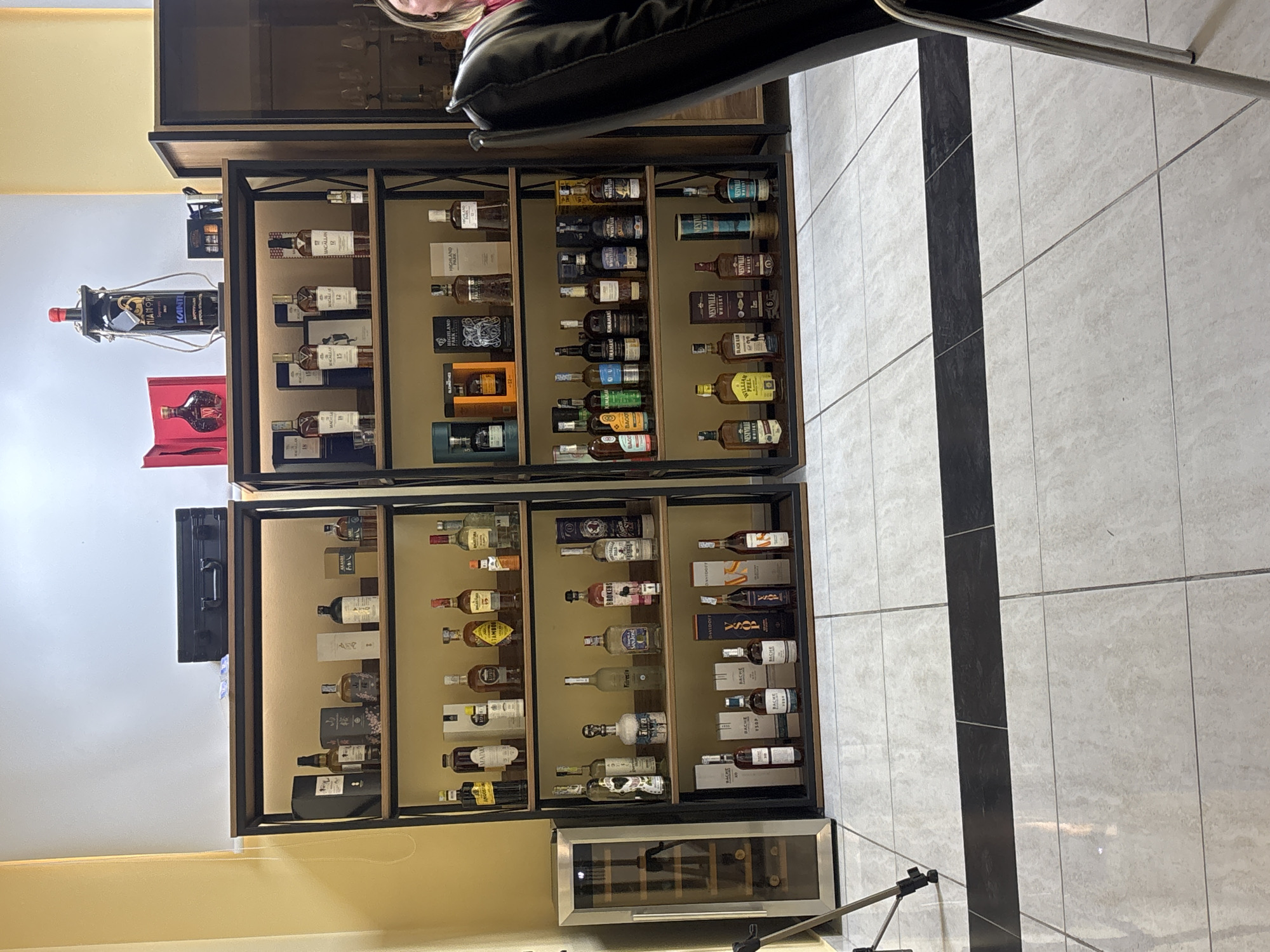 An indoor scene in Tbilisi, Georgia, features two tall, modern display shelves filled with an extensive collection of alcoholic beverages, predominantly whiskey, but also gin, brandy, and a hanging bottle of wine. The shelves are constructed with dark frames and light wooden back panels, and are internally lit, highlighting the bottles within.
The upper shelf on the left side displays several bottles of Macallan (including 12 and 15 year expressions) and Highland Park whiskies. The upper right section showcases a variety of whiskies such as Nestville Whisky, William Peel, and Black Ram, alongside Brokers Gin and Kinahan's Irish Whiskey. Below this, brands like Bago and San Jose are visible.
The lower shelf continues the collection, with more diverse spirits. Bottles from Bache (including VSOP), Davidoff, and Angostura are arranged neatly. A small wine cooler/fridge is positioned at the bottom, suggesting a curated beverage area.
On the left wall, a bottle of Kanti Niamori wine is suspended. Below it, a small bottle rests in a red box. A black case is mounted on the wall above the shelves.
A person is partially visible in the upper right corner, seated in a black chair. The floor is tiled with large light grey squares, featuring a striking dark grey vertical stripe running down the center. A photographic tripod or stand is visible at the very bottom right. The walls are a mix of light yellow/beige and white. The overall impression is a well-organized and sophisticated display of a personal spirits collection.