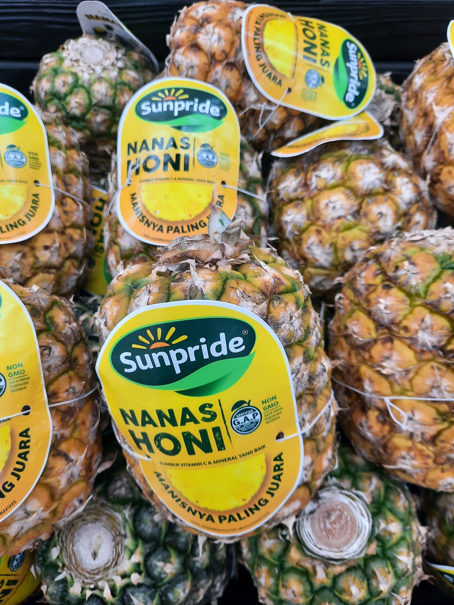 The image displays a close-up, overhead view of multiple ripe pineapples for sale, likely in an outdoor market or grocery store in Babakan Madang, Indonesia. The pineapples are characterized by their spiky green crowns and segmented, golden-brown skin. Several of the pineapples are adorned with bright yellow, oval-shaped labels that feature branding and product information. The dominant label text visible is "Sunpride NANAS HONI," indicating the brand and type of pineapple. Other text on the labels, though partially obscured, includes phrases like "SUMBER VITAMIN C & MINERAL YANG BAIK" (Source of Vitamin C & Good Minerals) and "MANISNYA PALING JUARA" (Sweetest Champion). Some labels also bear certifications such as "NON GMO" and "CERTIFIED G.A.P. AGRICULTURAL PRACTICES." The lighting suggests an ambient, possibly natural light source. The overall impression is one of fresh produce ready for purchase.