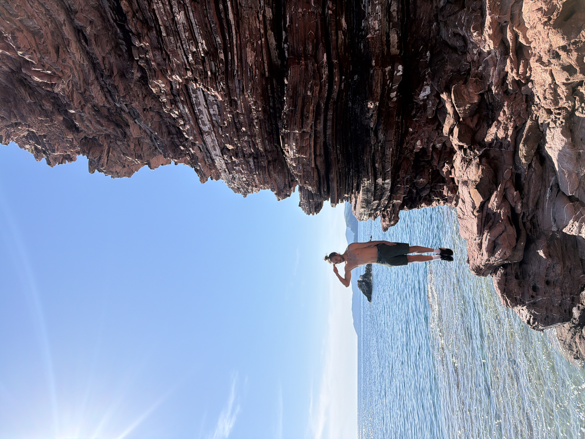 A rotated, wide-angle outdoor shot depicts a man standing on the edge of a rocky cliff overlooking the sea. The top part of the frame is occupied by a rocky overhang with layered, reddish-brown rock formations, suggestive of sedimentary strata. The lower portion shows a clear, bright blue sky and the shimmering surface of the sea. The man, who appears to be tanned, is wearing shorts and shoes and has his hair tied in a bun. He is seen making a gesture with one hand near his head. In the background, a rock formation rises from the sea, suggesting a coastal environment. The location is likely Sveti Stefan, Montenegro, based on the coastal features and rock formations.