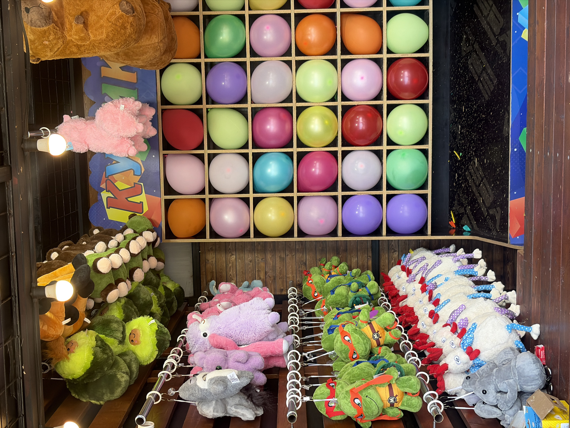 The image displays a vibrant carnival game booth, likely featuring a balloon-popping challenge. The central focus is a large wooden grid filled with numerous inflated balloons, meticulously arranged in rows and columns. These balloons come in a wide spectrum of colors, including red, orange, yellow, various shades of green, light blue, dark blue, purple, pink, and white.
Below the colorful balloon display, an extensive collection of plush toys is neatly arranged on wooden shelves, serving as potential prizes. On the left side, a large brown plush bear hangs partially visible at the top, followed by a bright pink fluffy animal illuminated by a light bulb. Further down, green avocado-shaped stuffed toys are stacked. The central shelves hold rows of pink and purple furry animals, alongside grey plush toys. To the right, numerous green Ninja Turtle-like characters, each with a red or orange mask and a brown shell-like backpack, are lined up. On the far right, a series of white chicken-shaped plush toys with red combs and blue or purple polka-dotted legs are visible.
The entire setup is brightly lit by artificial light bulbs, one of which is prominently visible on the left side, casting a glow on the prizes. The background features various elements, including a dark wall with faint patterns behind the balloons and abstract colorful shapes on a blue background on the right.
Visible text includes the words "Sweet Toys" on a tag attached to one of the pink plush animals. Partial letters "KY" are seen on a colorful background to the left of the balloon grid, and the word "GROUP" is faintly visible on the dark wall to the right. The scene evokes the lively atmosphere of an amusement park or fair.