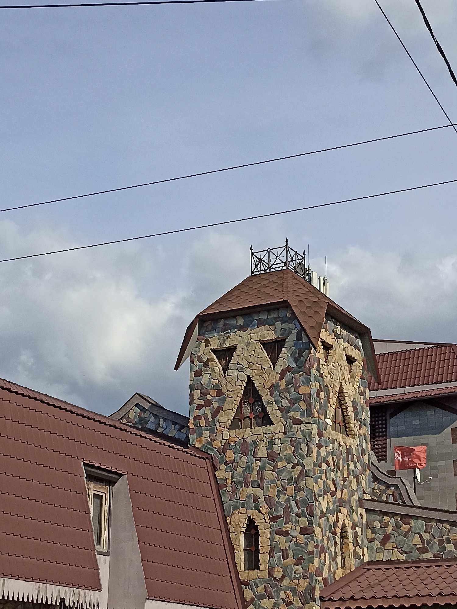 The image shows a view of a building in Mochische, Russia, featuring a prominent stone tower with triangular windows. The tower is constructed from a mosaic of multicolored stones, ranging from beige and brown to grey and hints of blue. The roof of the tower is covered in brown shingles and topped with a decorative metal railing. Adjacent to the tower are other parts of the building, also with brown shingled roofs. One roof section has a small window with a wooden frame. A red flag with a Cyrillic inscription is visible on a pole attached to another part of the building. The sky above is partly cloudy, with power lines crossing the frame horizontally. The scene appears to be an exterior shot of a building, possibly a house or a commercial establishment. There are no people visible in the image.