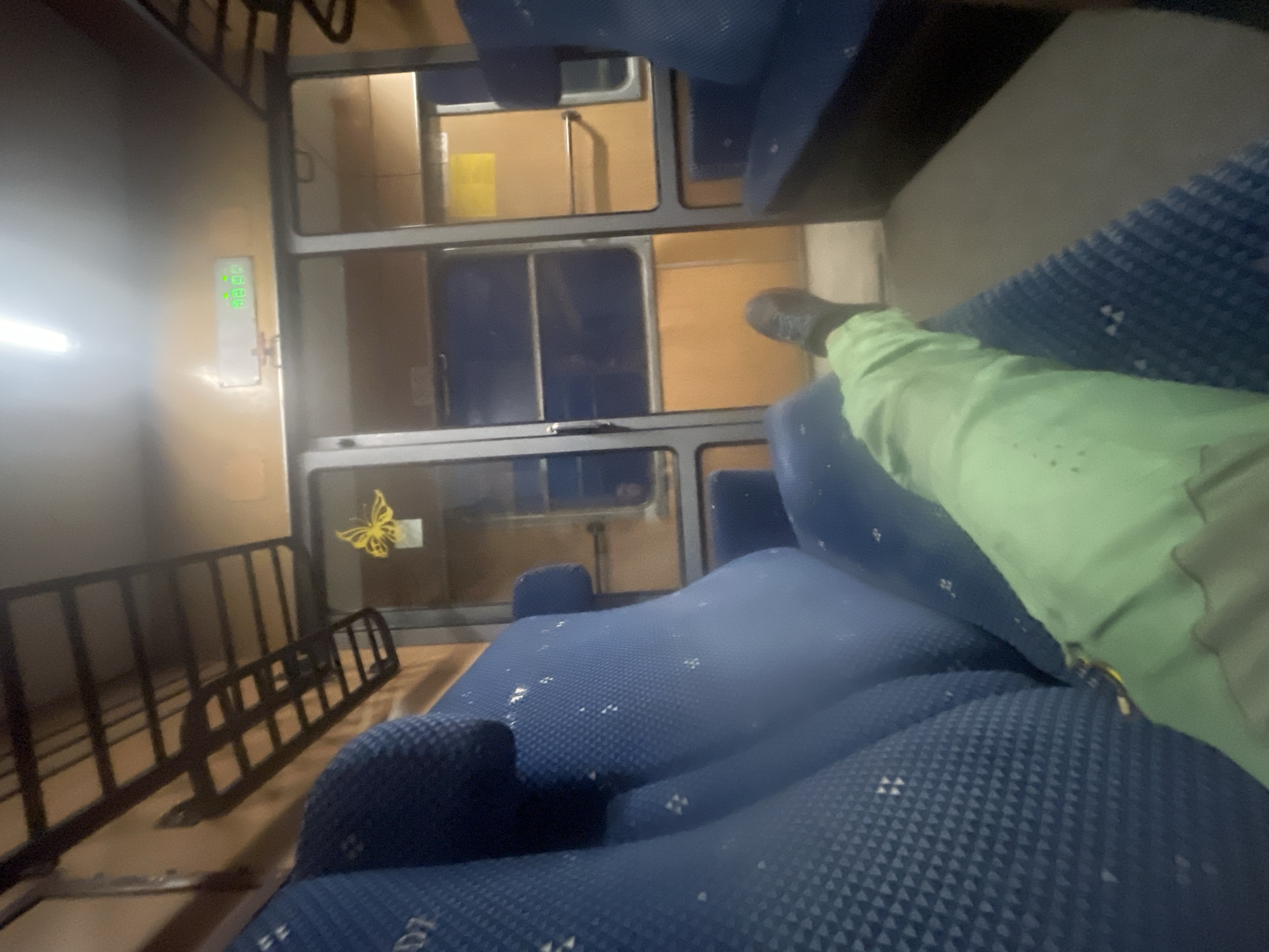 The image shows the interior of a train carriage. A person's legs, wearing light green trousers and a dark shoe, are visible extending into the frame, resting on blue seats. The seats appear to be upholstered with a textured fabric. In the background, there is a compartment with glass doors, revealing shelves and orange-colored walls. A yellow butterfly sticker is affixed to one of the glass panes. Above the compartment, a black metal luggage rack is installed. A digital display with green numerals is mounted on the wall, showing "19:19". The train carriage appears to be dimly lit with a fluorescent light fixture visible at the top of the image. The environment suggests a journey on a train, potentially within or traveling to Borovo, Bulgaria, though the location is not explicitly confirmed by visual cues within the image itself.