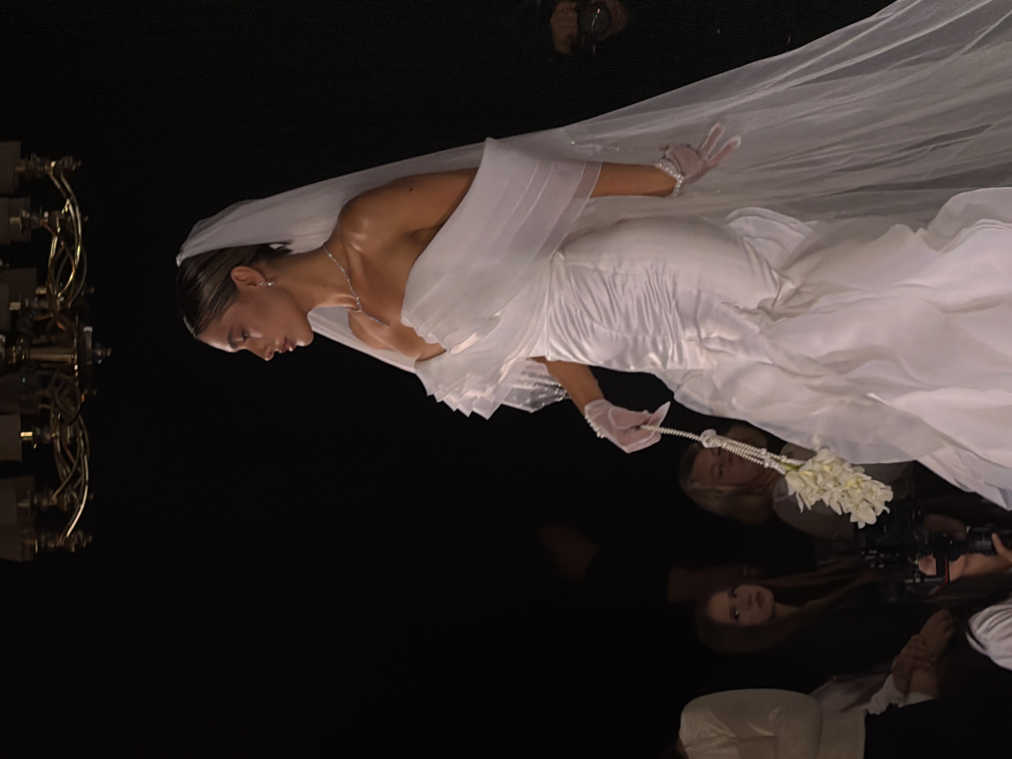 Here is a description of the image:
A model wearing a wedding dress and veil is photographed in an unspecified location in Unknown, Unknown. She is accessorized with gloves and is holding a bouquet of flowers. The scene includes a person with a camera and lighting equipment is visible. The overall environment is dark with light focused on the model.