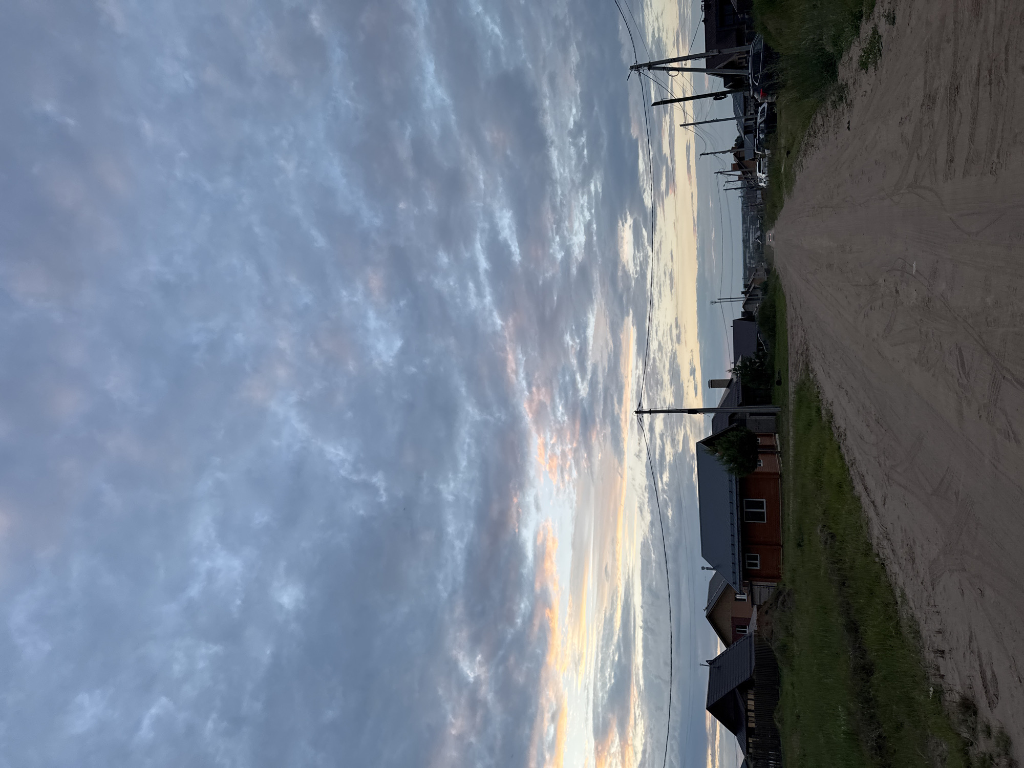 This image depicts a dirt road in a rural or suburban area, likely on the outskirts of Chaikovsky, Russia. The road is unpaved and shows tire tracks, suggesting vehicular traffic. On either side of the road, there are houses with dark roofs and wooden exteriors, typical of a dacha settlement or village. The vegetation appears to be grass and some trees, indicating a natural or semi-natural environment. In the background, there are more houses and power lines stretching along the road, further suggesting a residential area. The sky is filled with clouds, with hints of orange and pink hues near the horizon, indicating either sunrise or sunset. There are no people visible in the image. The overall impression is one of a quiet, undeveloped, or developing community.
