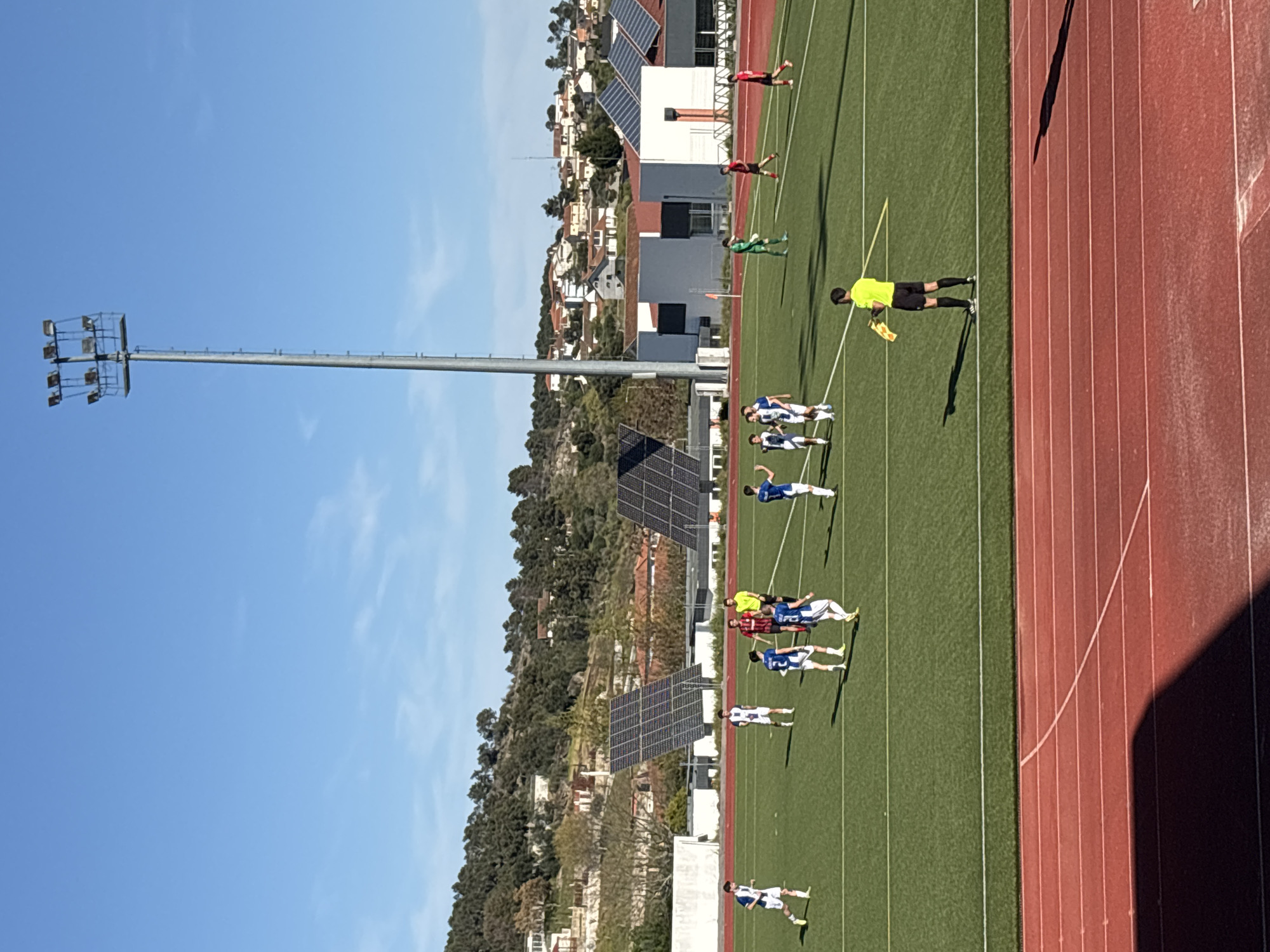 The image shows a soccer game in progress in Valpaços, Portugal. Players in blue and white uniforms are positioned on a green soccer field. A referee in a yellow shirt stands near the sideline. A red running track is adjacent to the field. A tall light pole stands near the field. The background includes buildings with solar panels and trees on a hill. The sky is blue.