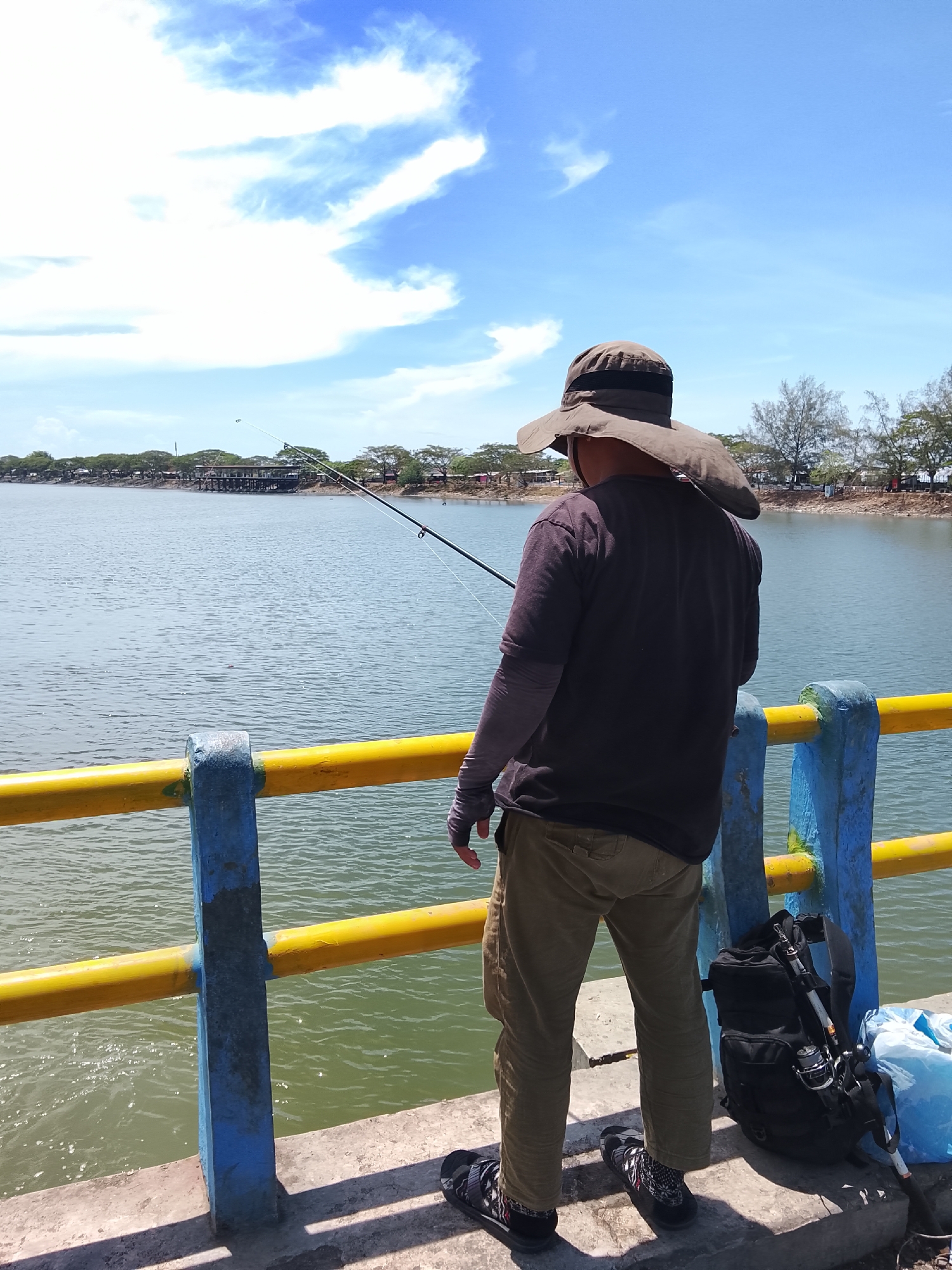 A person stands on a concrete pier, facing away from the camera and towards a body of water. The person is wearing a wide-brimmed hat, a dark t-shirt with long sleeves, khaki pants, and sandals. A fishing rod is held in the person's hands, and the line extends into the water. To the right of the person, a black backpack with a fishing rod attached is placed on the pier, along with a blue bag. The pier is lined with a yellow railing supported by blue posts. The water is calm and reflects the blue sky with scattered white clouds. In the distance, across the water, is a shoreline with trees and some buildings. The scene appears to be outdoors on a sunny day. The location is the reservoir in Lhokseumawe, Indonesia.