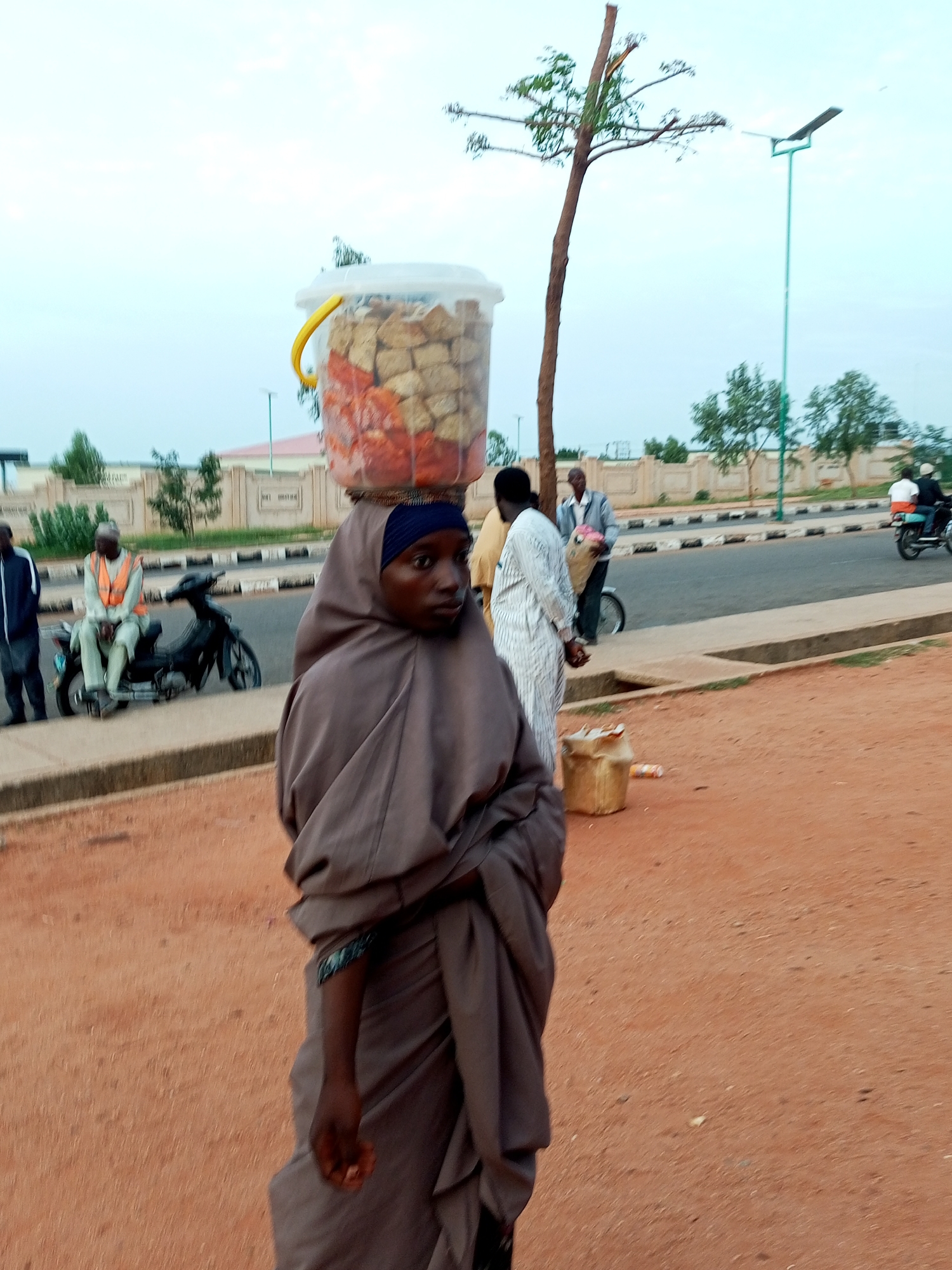 In Gombe, Nigeria, an outdoor scene captures a young woman carrying a large plastic bucket filled with what appears to be food items, balanced on her head. She wears a brown hijab and traditional attire. Behind her, a man sits on a motorcycle, wearing an orange safety vest, and another man stands beside him. In the background, several buildings with decorative walls and trees are visible, along with a solar-powered street light. The ground is covered with reddish-brown dirt. The overall atmosphere suggests a typical street scene in a bustling Nigerian city.