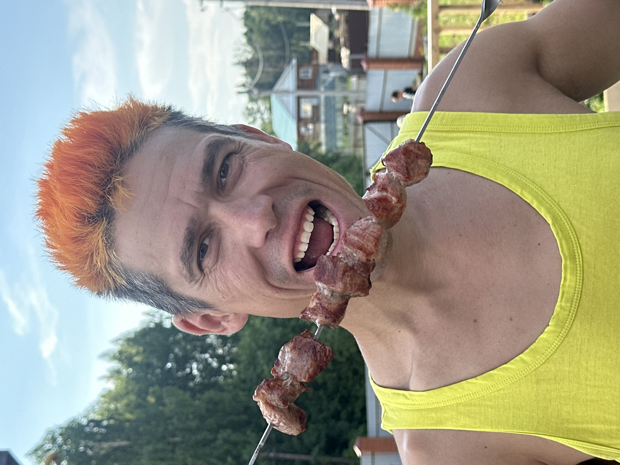 A middle-aged man with bright orange hair is holding a skewer of grilled meat up to his open mouth outdoors on a sunny day. He is wearing a yellow tank top. In the background, there are trees, a fence, and houses, suggesting a residential area. The man appears to be in good health and is smiling widely.